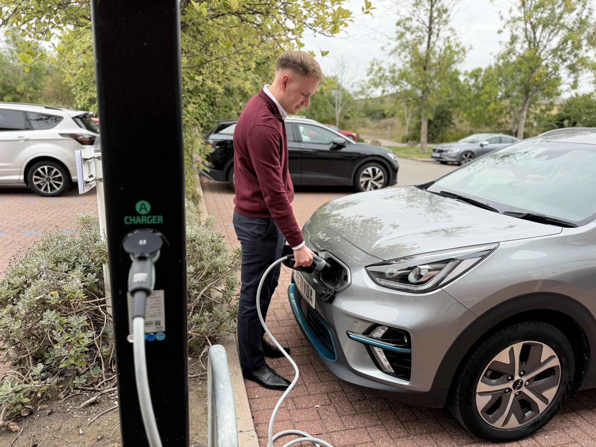 Man charging a gray electric car at a charging station in a parking lot.