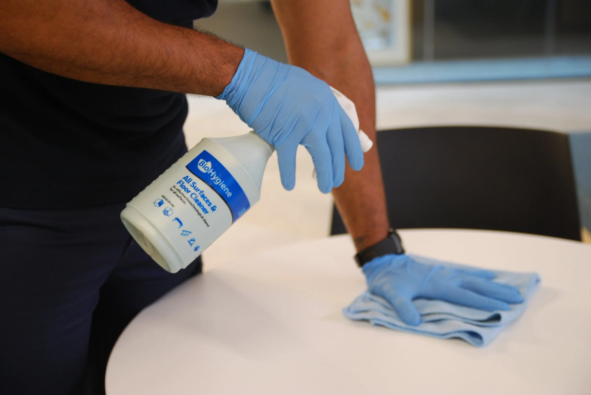 A person wearing black gloves cleaning a white table with a blue spray bottle and a cloth.