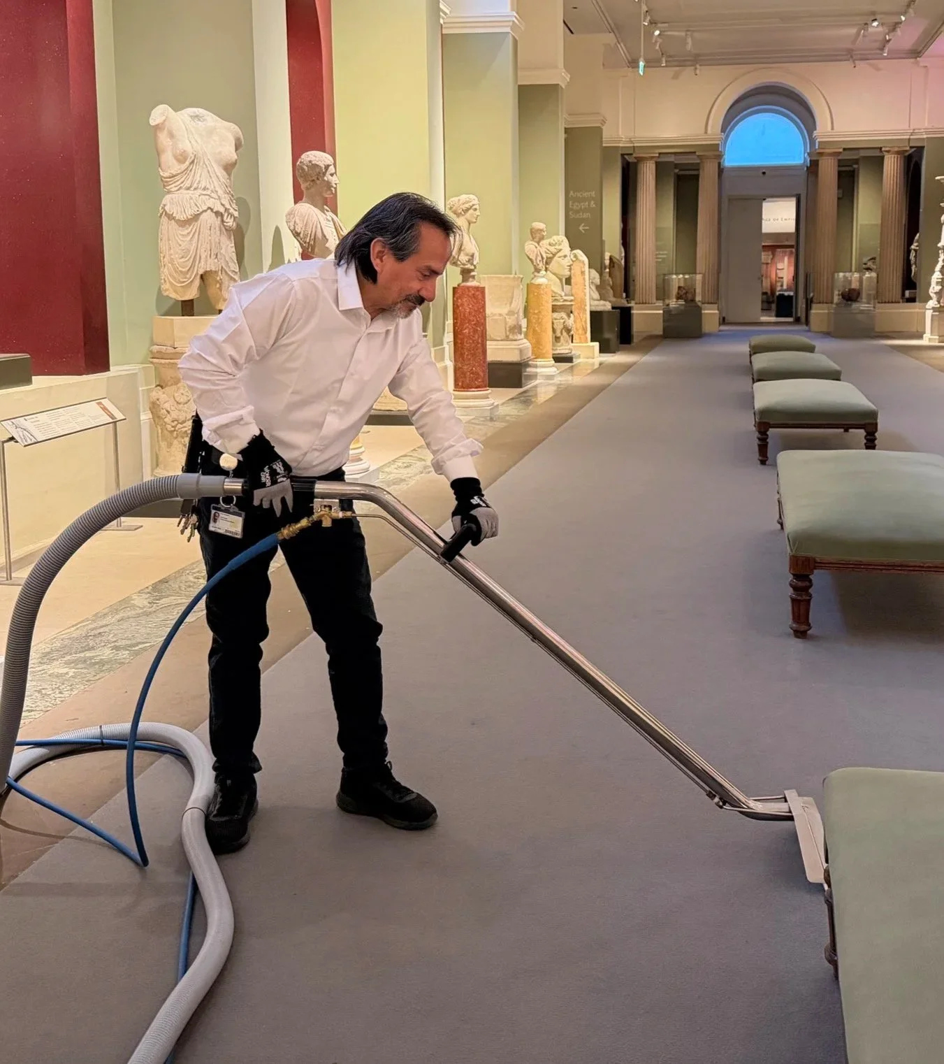 A man in a white shirt and black gloves vacuuming a carpet in a museum hallway with statues and columns.