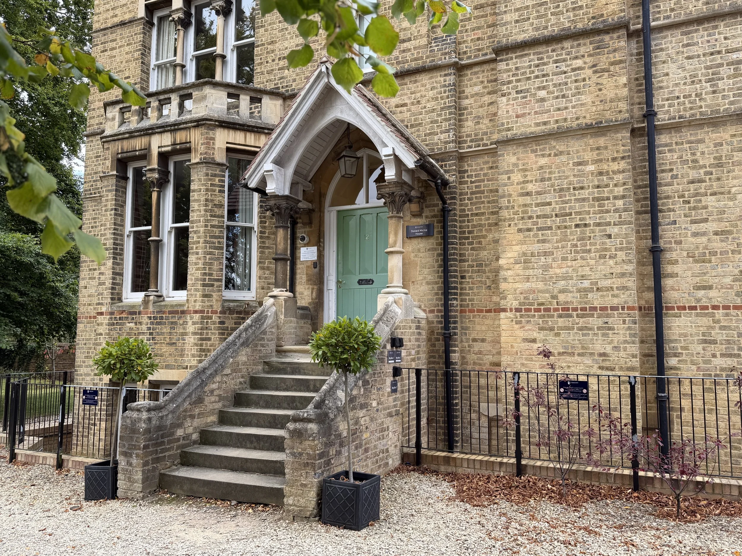 A historic brick building with a green front door, arched porch, stone steps, and decorative columns. There are small trees in planters on either side of the stairs, and a gravel path in front.