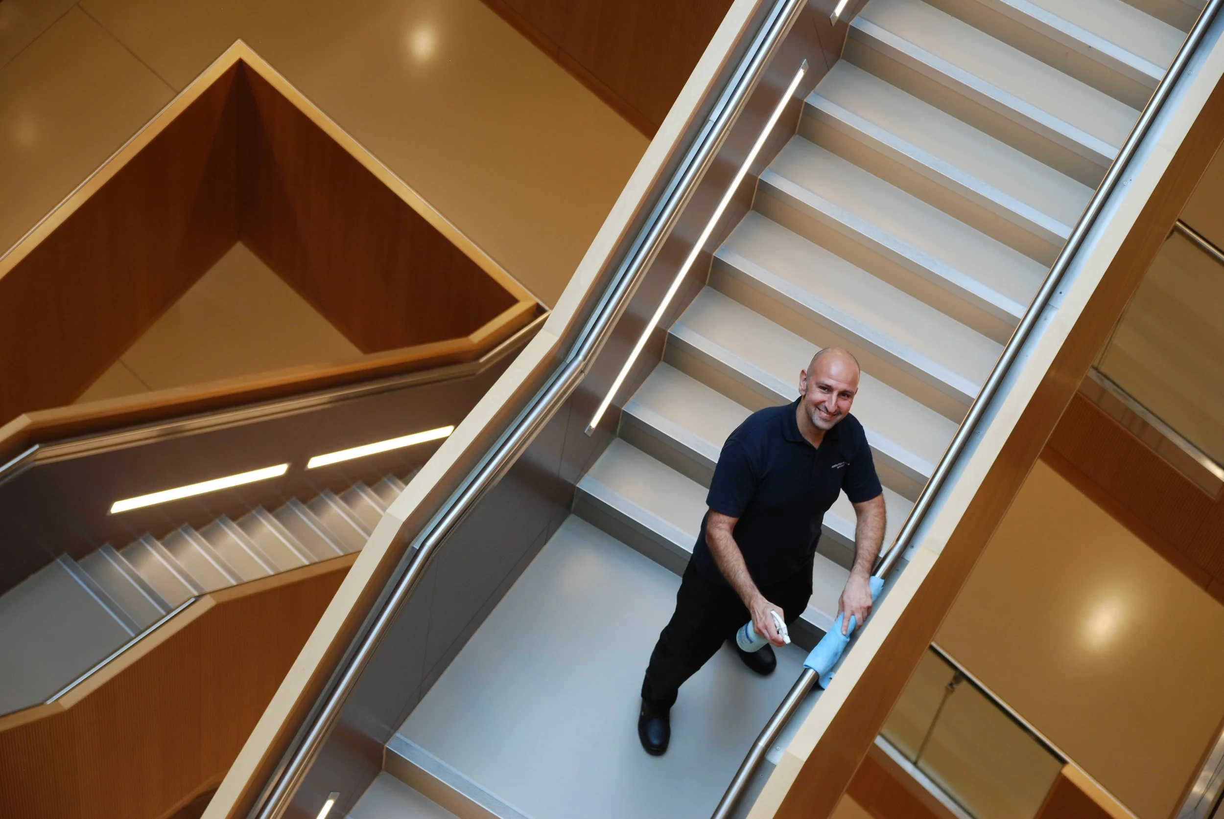 A smiling man standing on an elevator staircase, looking up at the camera, holding cleaning supplies.