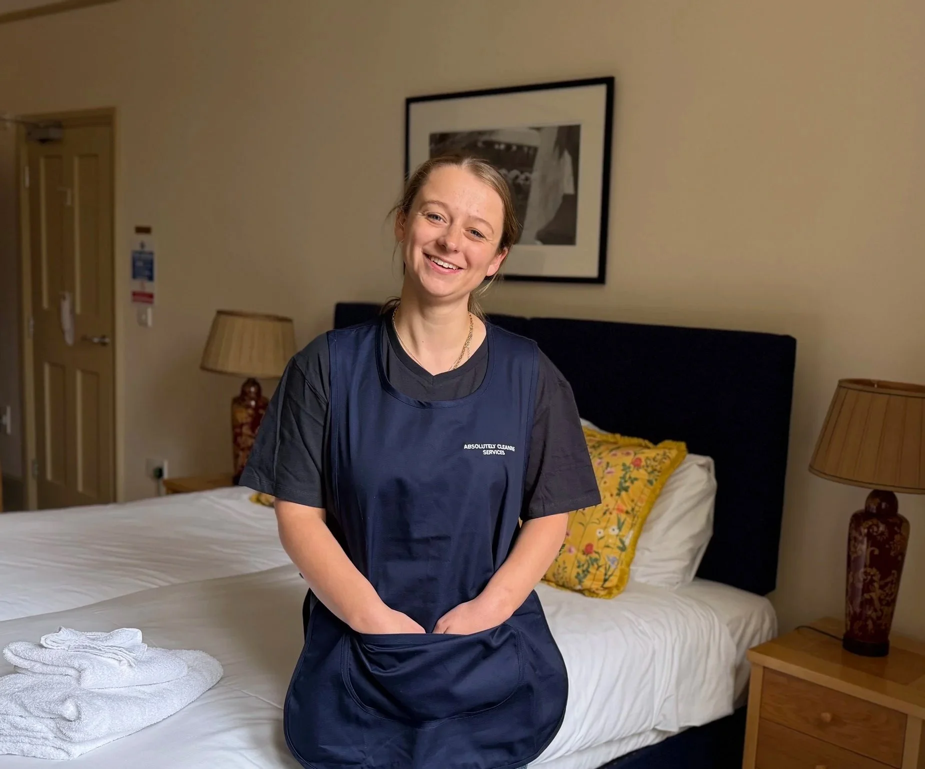 A woman in housekeeping uniform smiling in a hotel room, with a bed and bedside tables.