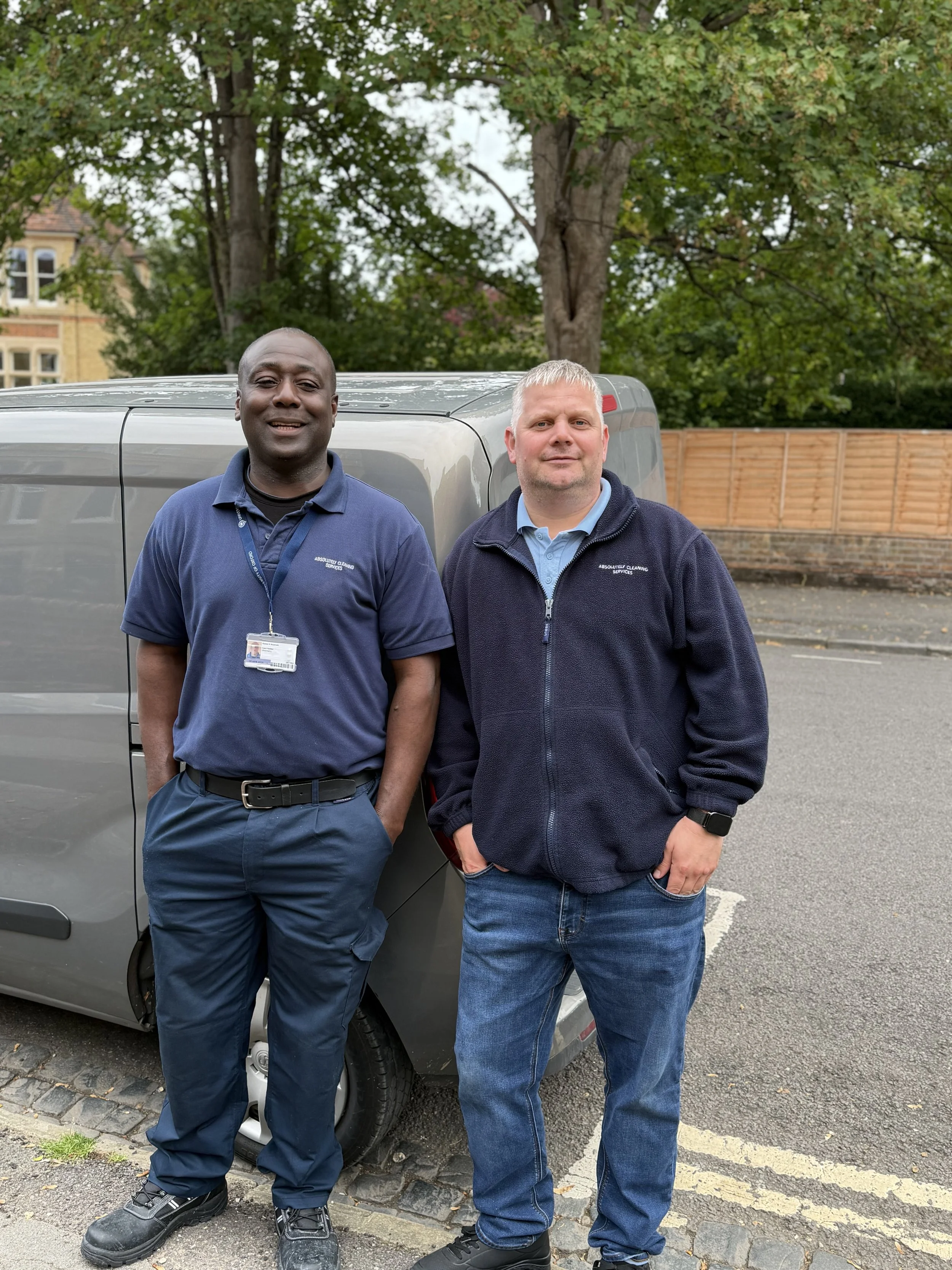 Two men standing in front of a gray van outdoors, with trees and a wooden fence in the background.