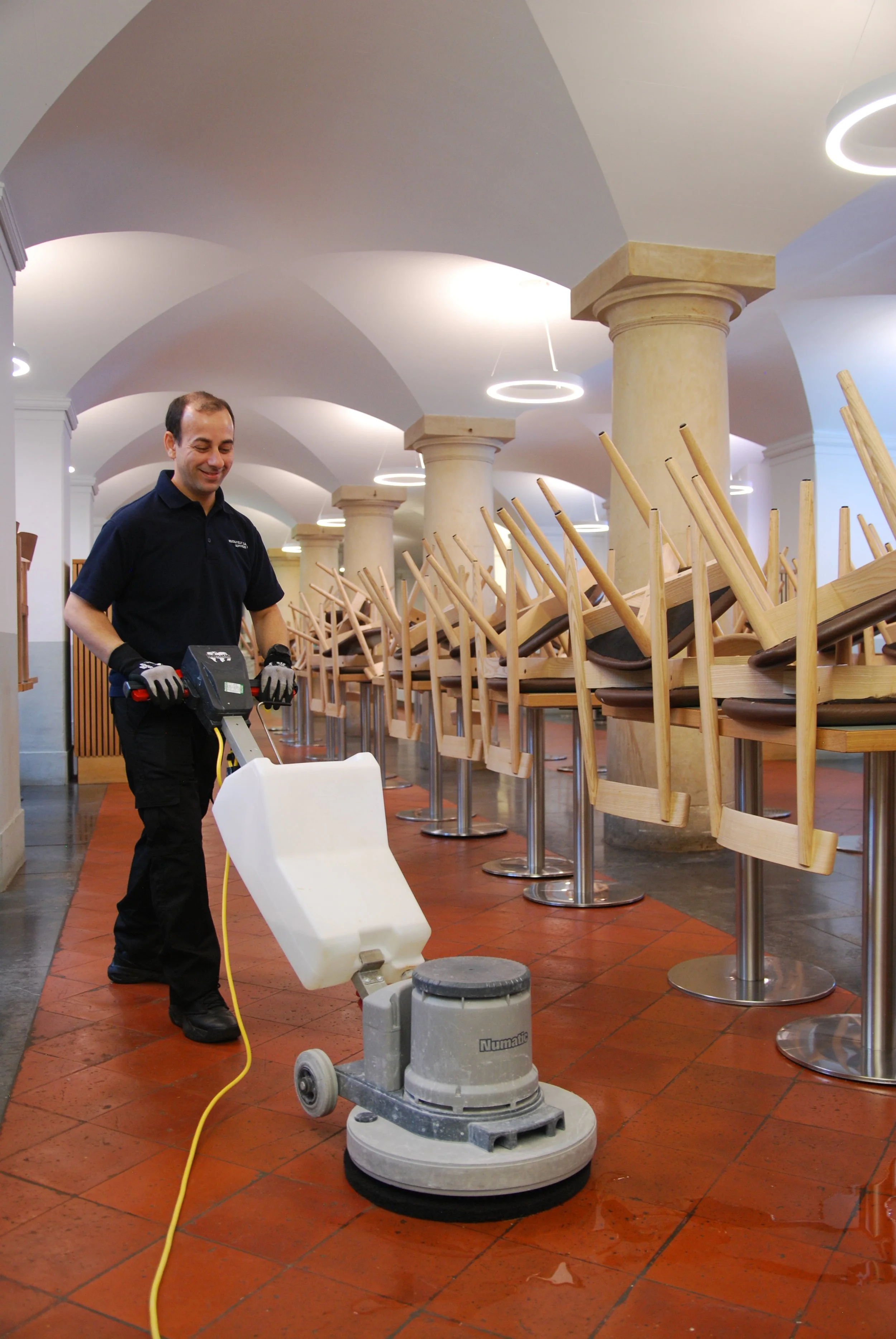 A man cleaning the floor with a floor buffer sander in a dining area with overturned chairs stacked on tables.