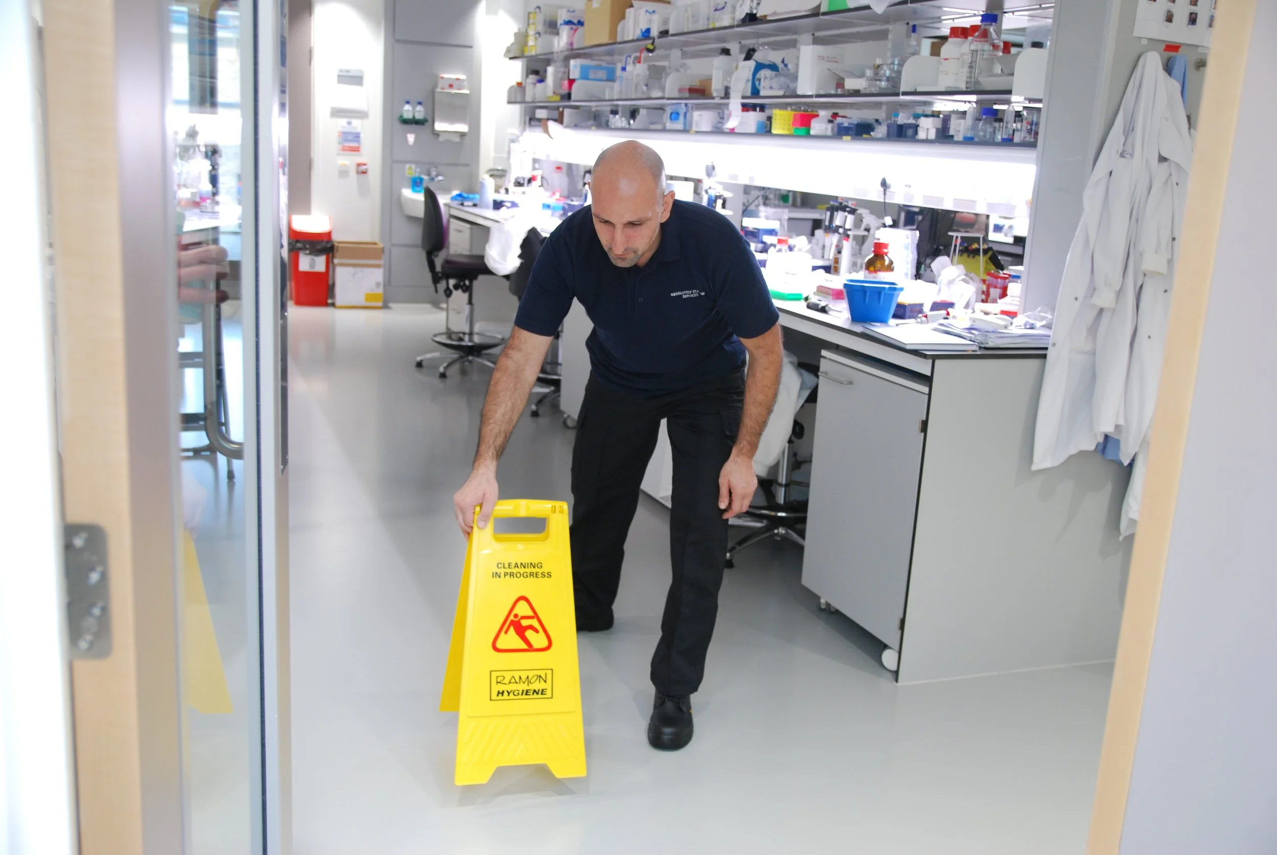 A man in a laboratory environments carefully places a yellow 'Cleaning in Progress' caution sign on the floor.