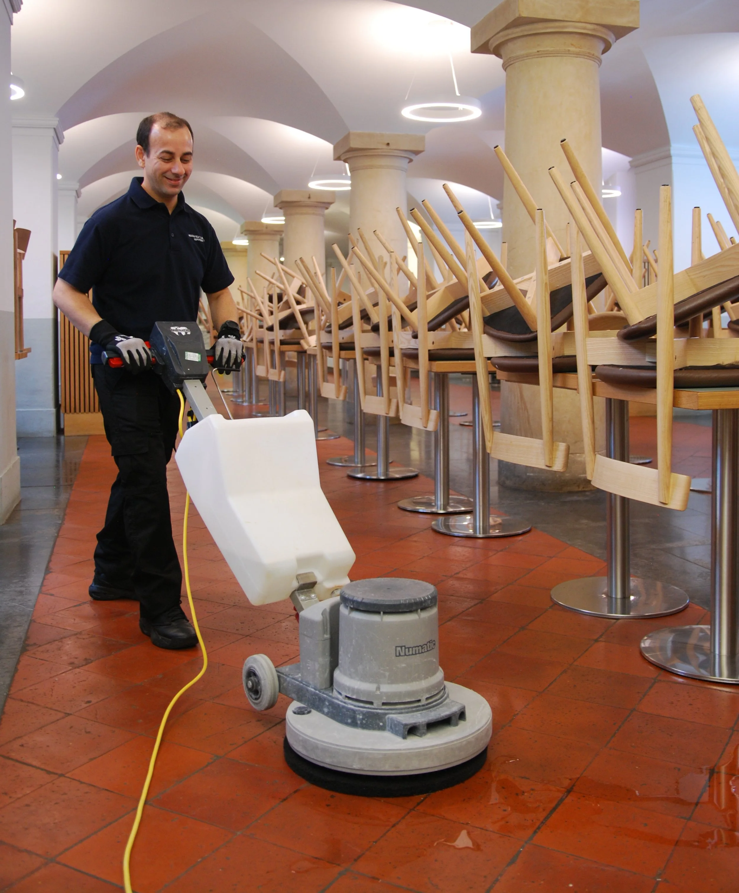 A man is using a floor polishing machine in an indoor restaurant or cafe with chairs stacked upside down on tables.
