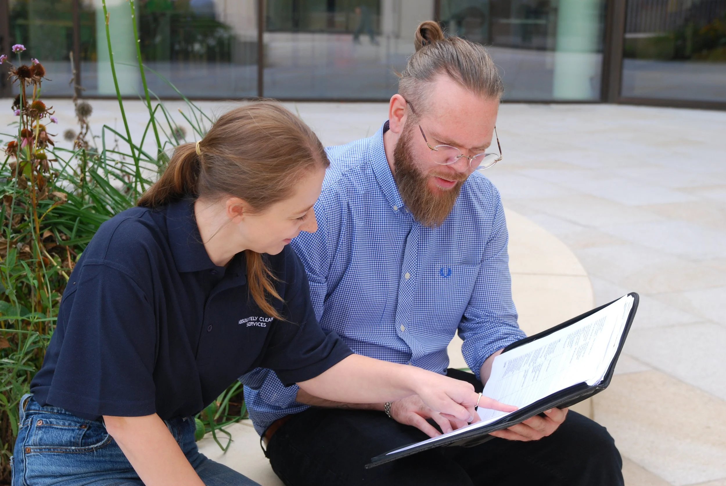 A man with a beard and glasses wearing a blue shirt sitting on a bench and a woman with brown hair wearing a dark blue shirt, sitting beside him, looking and pointing at a document or notebook they are examining together outdoors.