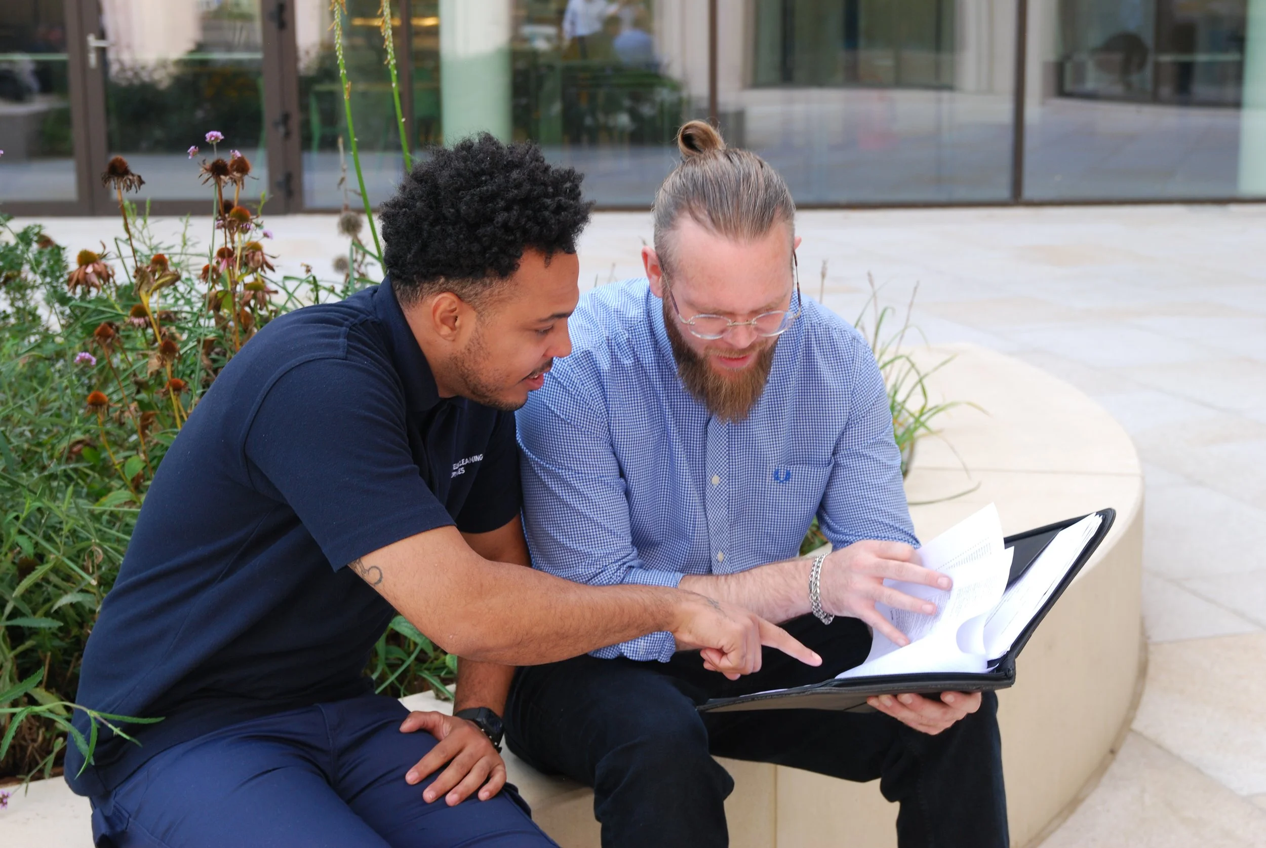 Two men sitting on a bench outside, looking at a binder or portfolio, engaged in a discussion.