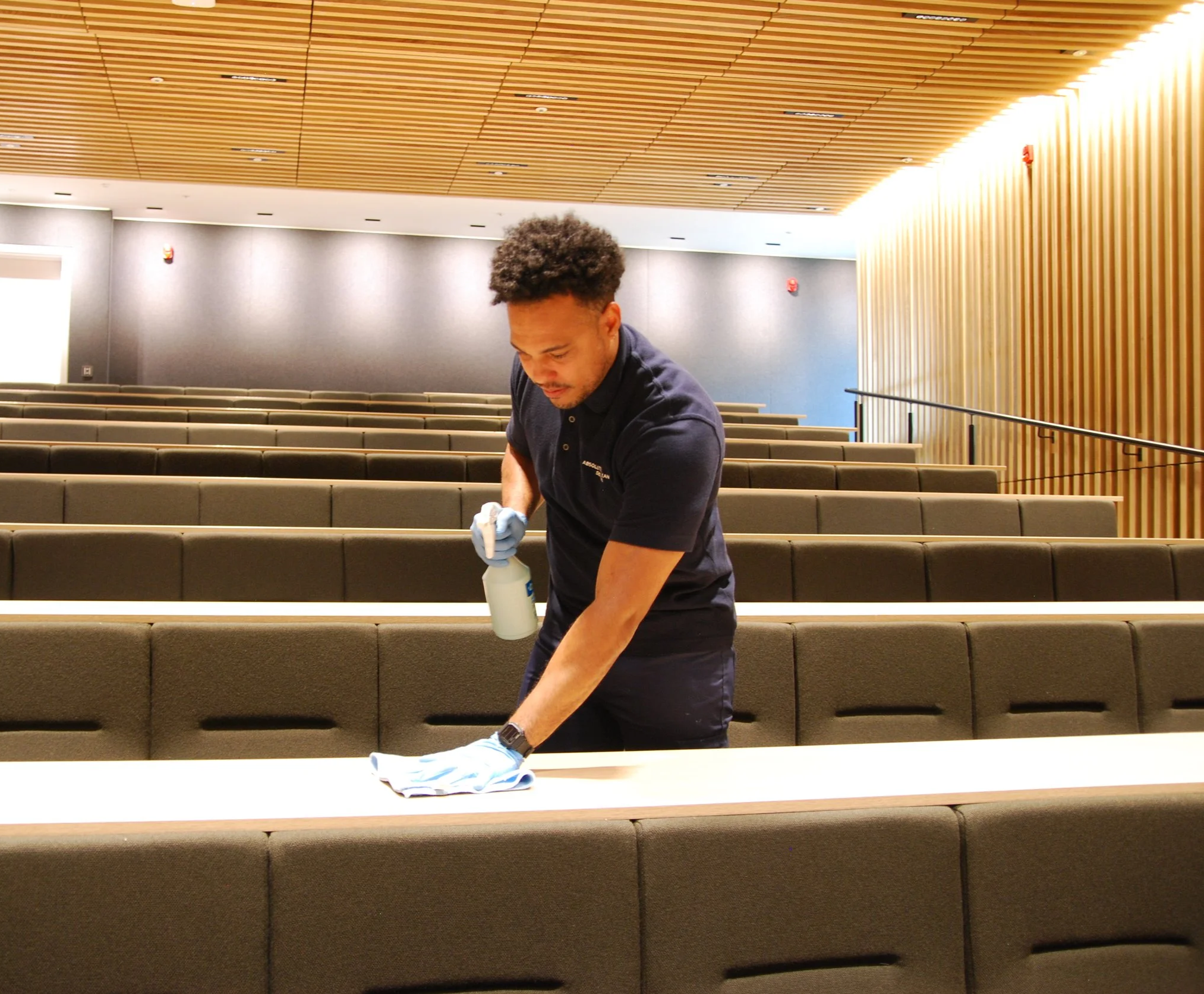 A man cleaning a conference room table with a spray bottle and cloth, wearing gloves.