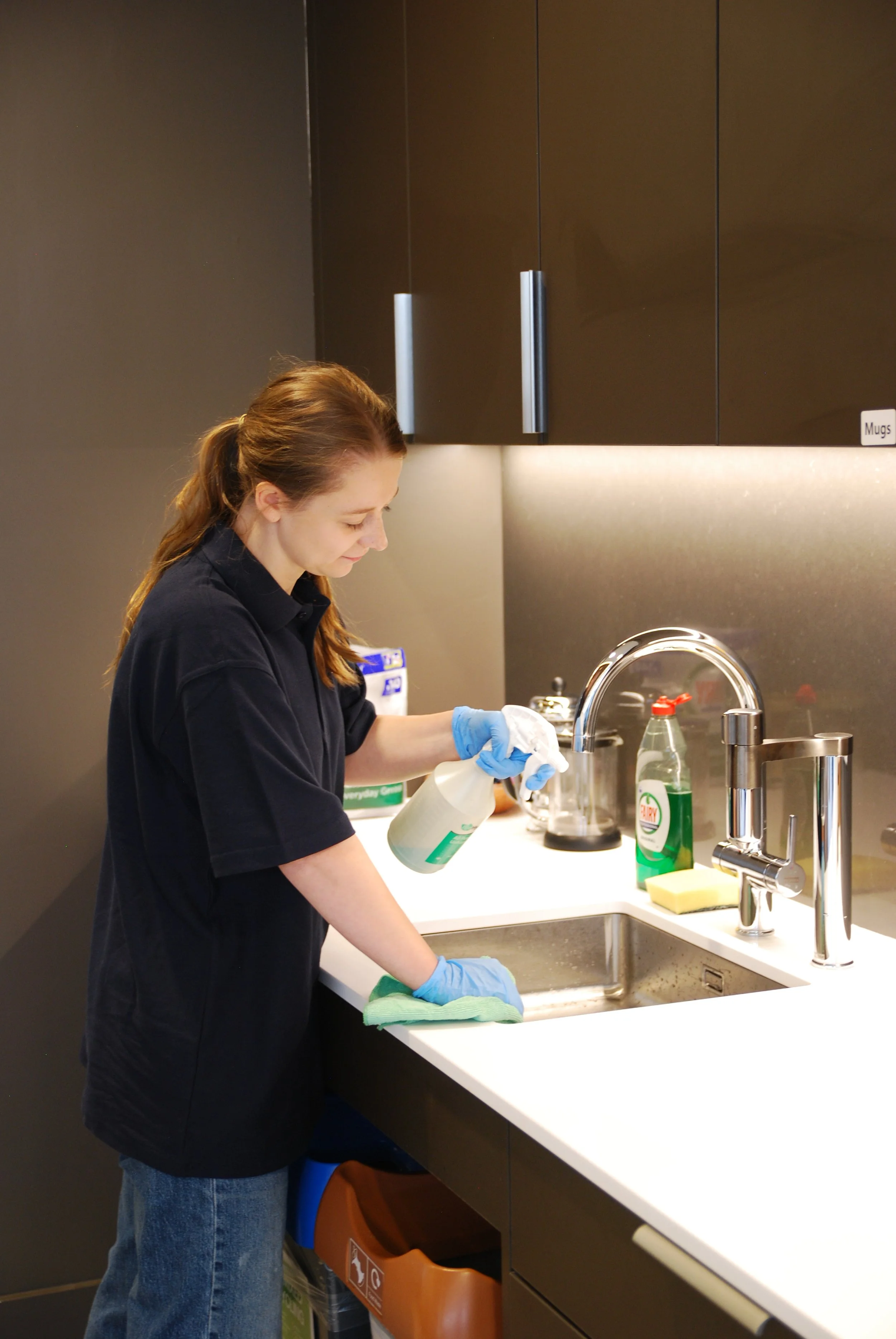 A woman wearing a black uniform and blue gloves is cleaning a kitchen sink with a spray bottle and a green cloth.