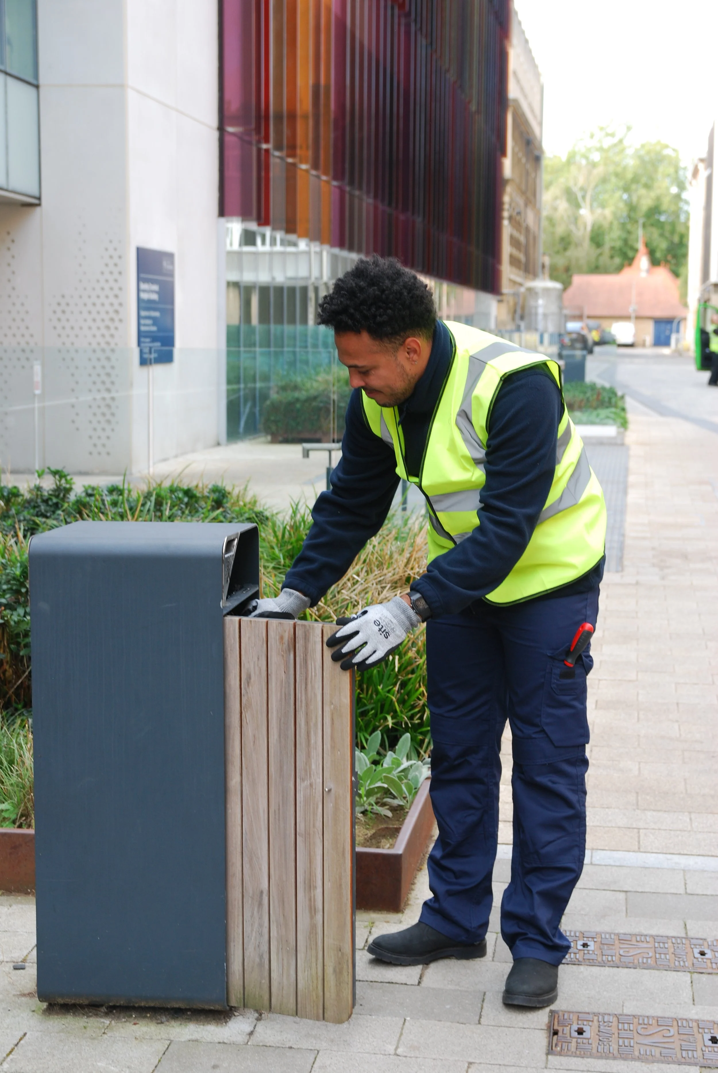 A maintenance worker in a high-visibility vest collects trash from a wooden and metal trash bin on a sidewalk in an urban area.