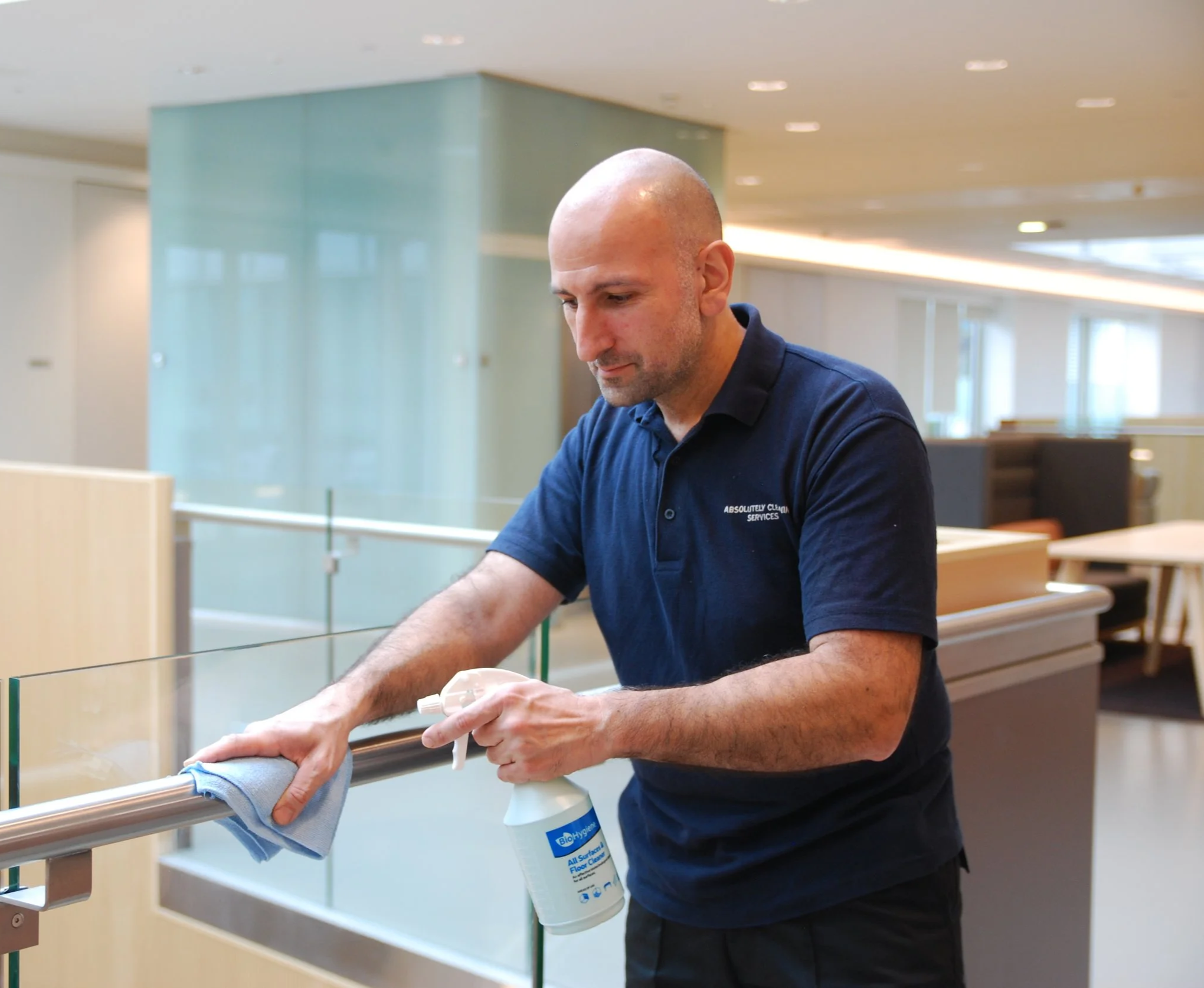 Man cleaning a glass railing with a spray bottle and cloth in a modern indoor space.