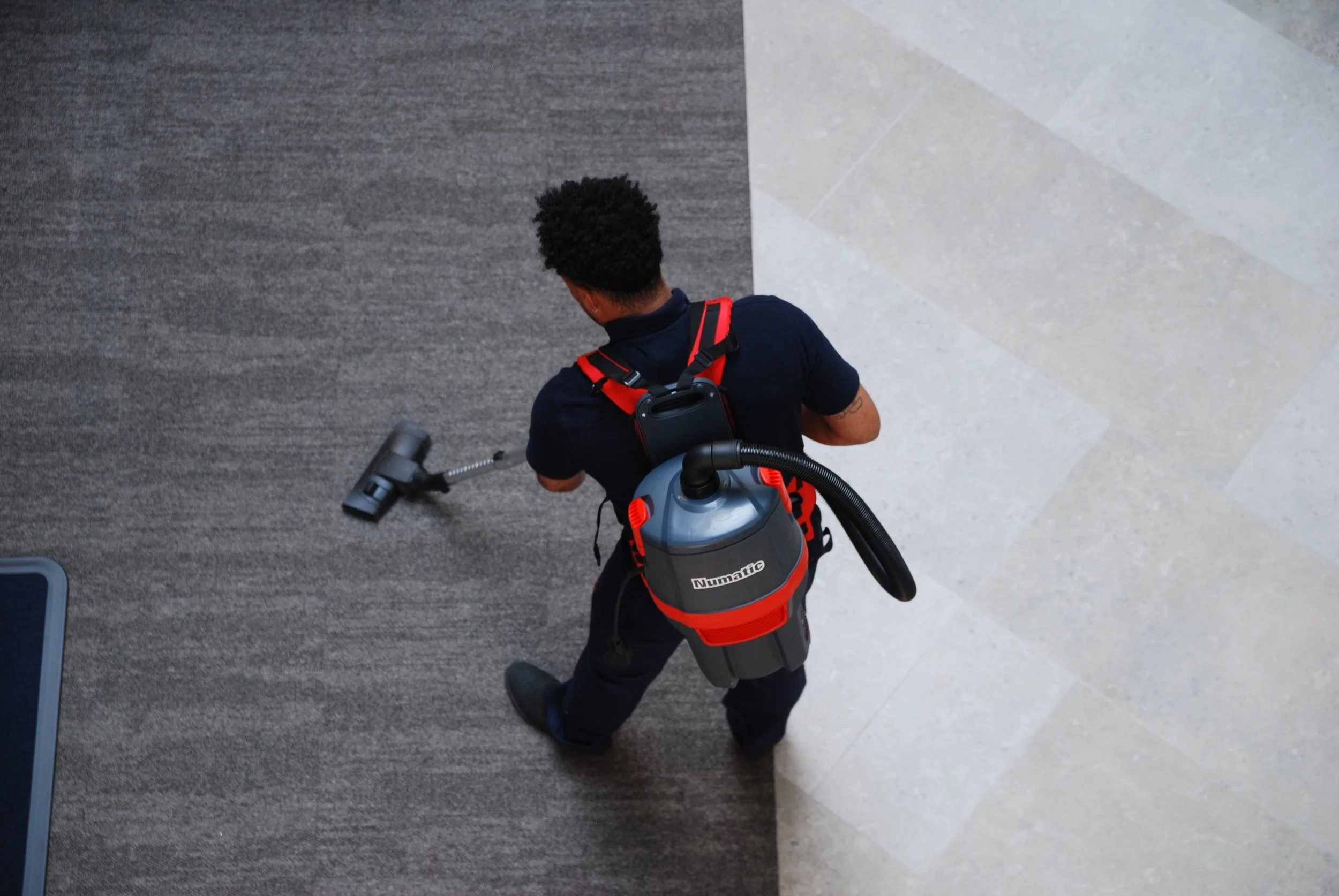 A man wearing a black uniform is vacuuming a carpeted floor with a Numatic brand vacuum cleaner, with a gray and black body and red accents, in a modern interior space.