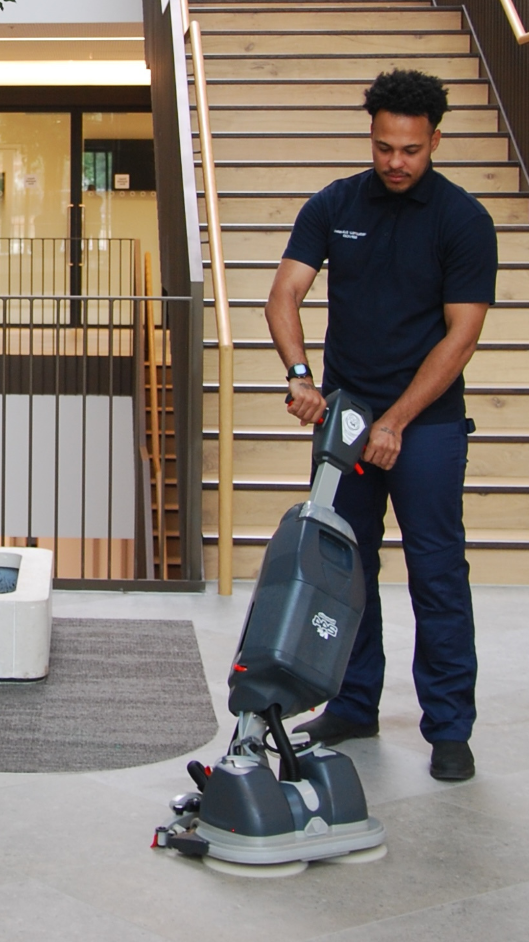A maintenance worker operating a floor cleaning machine inside a building with wooden stairs and railing in the background.