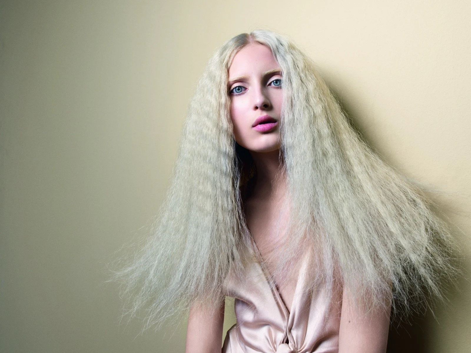 A woman with long, crimped platinum blonde hair and blue eyes, wearing a satin slip dress, standing against a plain beige wall.