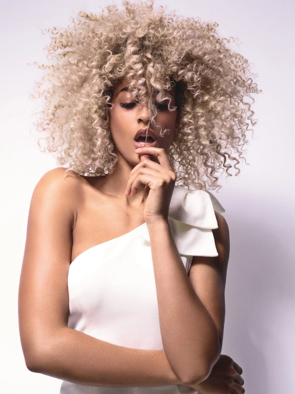 A woman with curly blonde hair in a fluffy style, wearing a white dress with an elaborate shoulder ruffle, posing with her eyes closed and finger touching her lips against a plain white background.