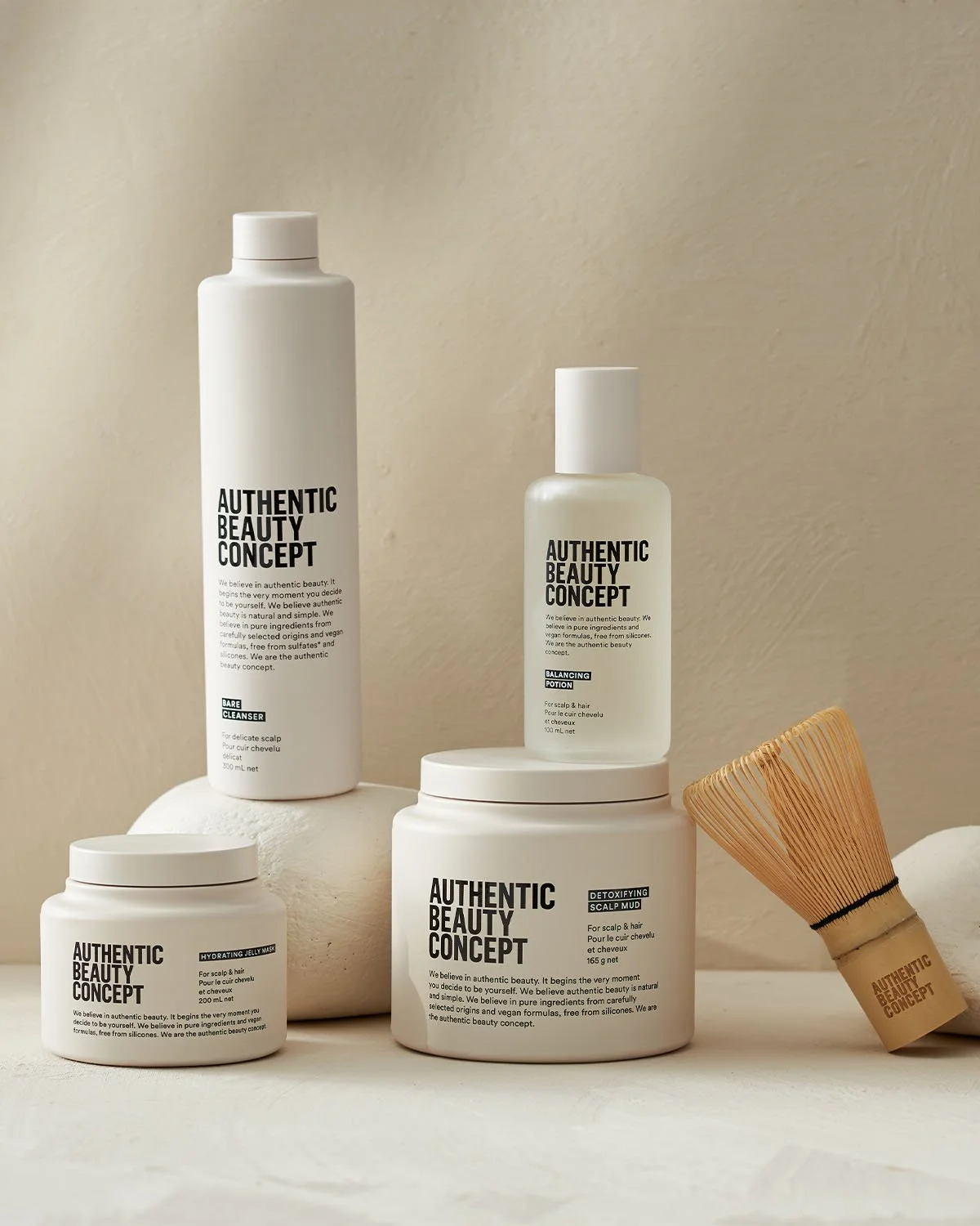 Collection of skincare and haircare products with minimalist white packaging, including bottles and jars, alongside a wooden scalp massager in a neutral setting.