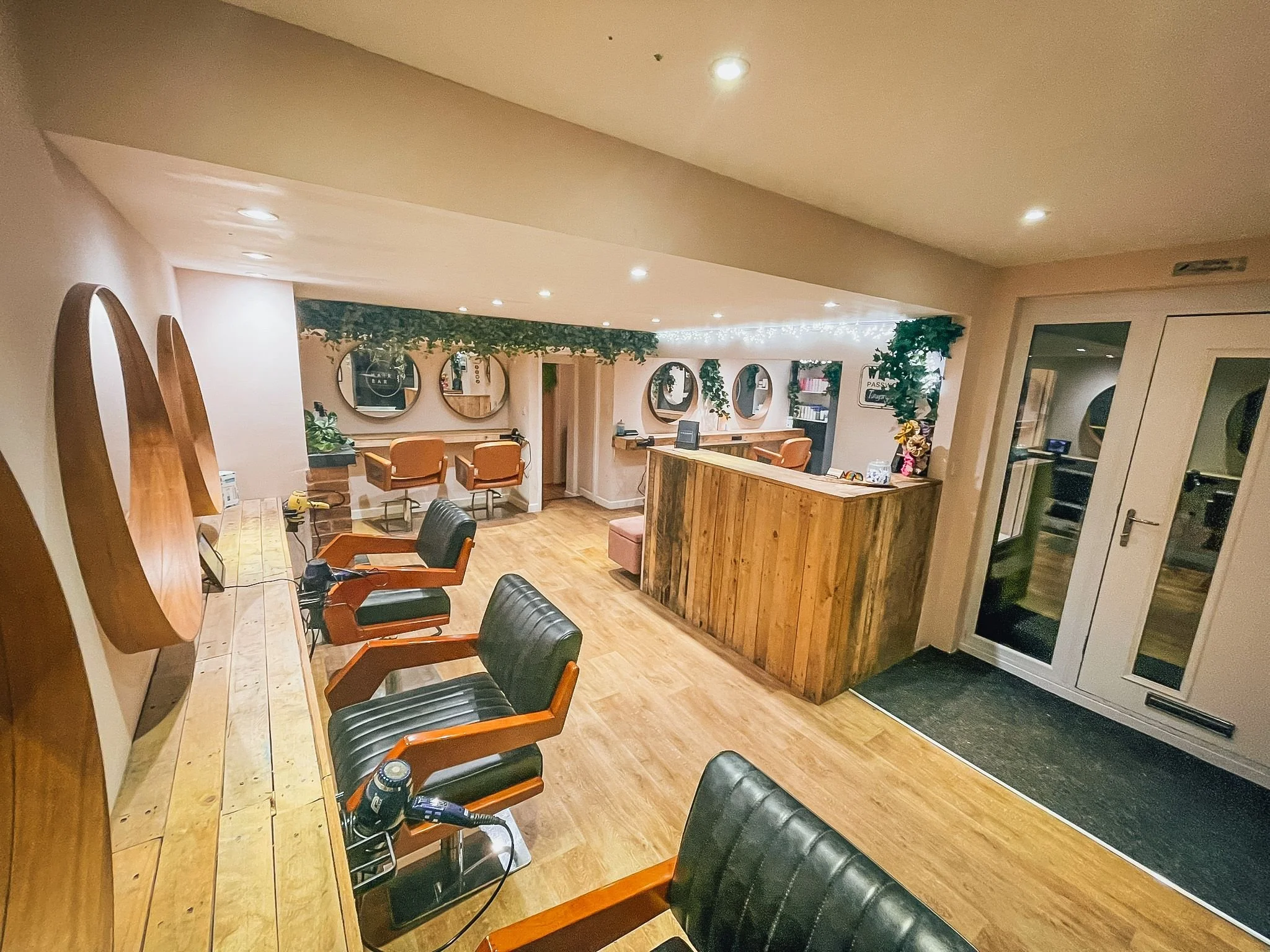 Interior of a hair salon or barbershop with chairs, mirrors, and a reception area decorated with plants and string lights.