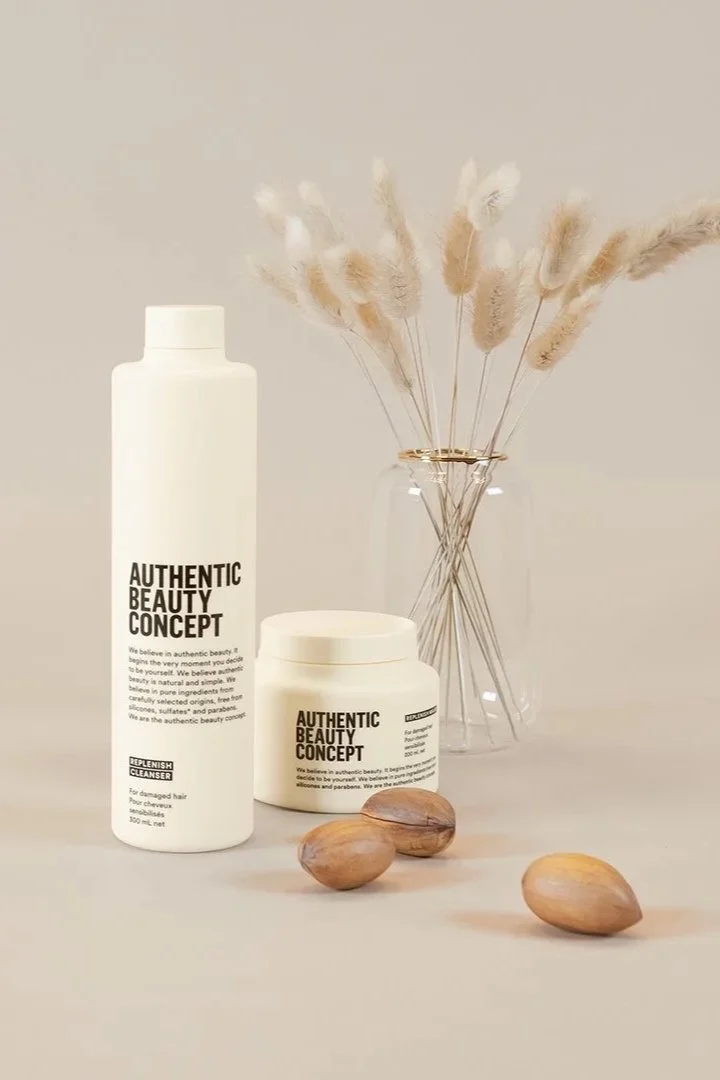 A minimalist arrangement of beauty and skincare products, including a tall white bottle, a shorter jar, and three almonds, with a clear glass vase holding beige pampas grass, all set against a neutral background.