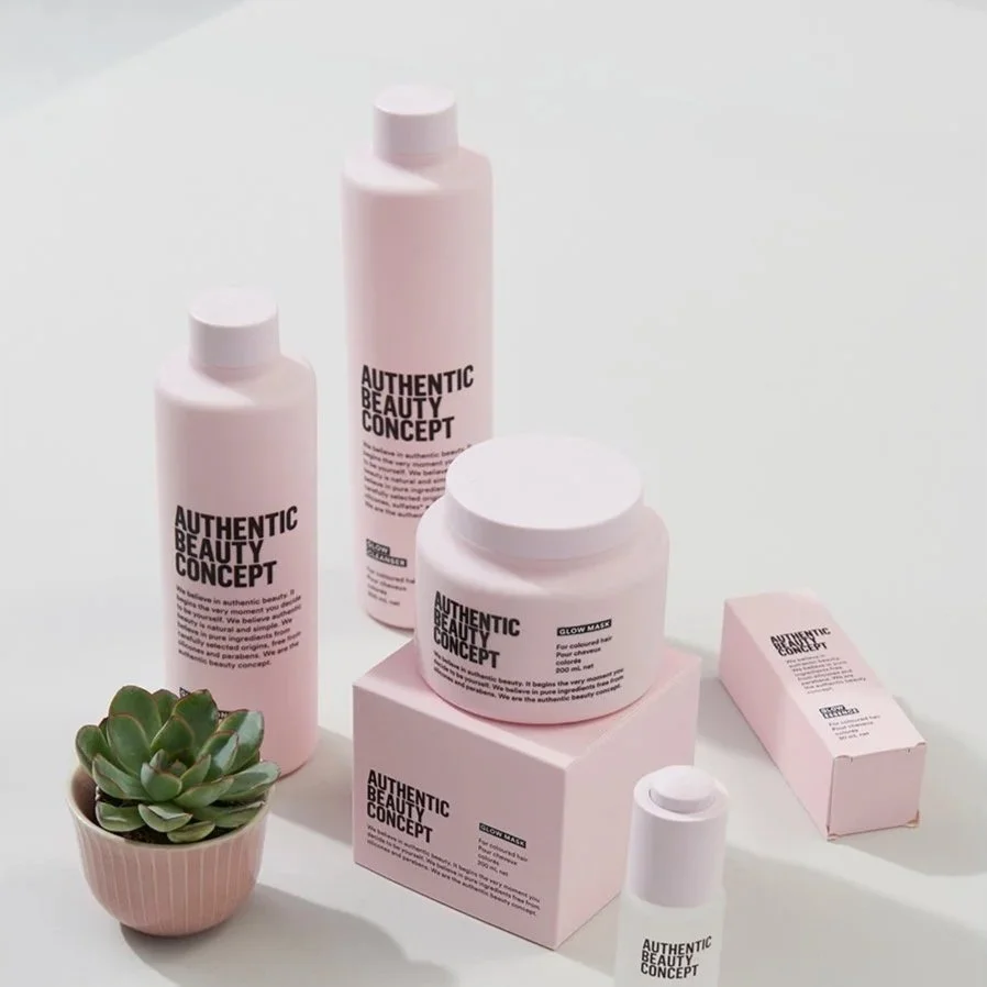 Collection of skincare products from Authentic Beauty Concept, including bottles, jars, a soap bar, and a small potted succulent plant, arranged on pink boxes and a white surface.