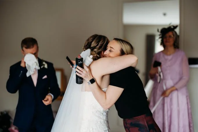 Two women embrace warmly at a wedding, with one in a wedding dress and veil and the other in a black shirt, hugging. In the background, a man in a tuxedo wipes his eyes with a tissue, and a woman in a pink dress holds a smartphone.