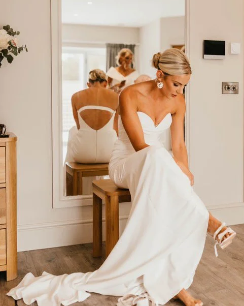 Bride in a white wedding dress sitting on a small stool, adjusting her shoe in a room with a mirror and her reflection visible.