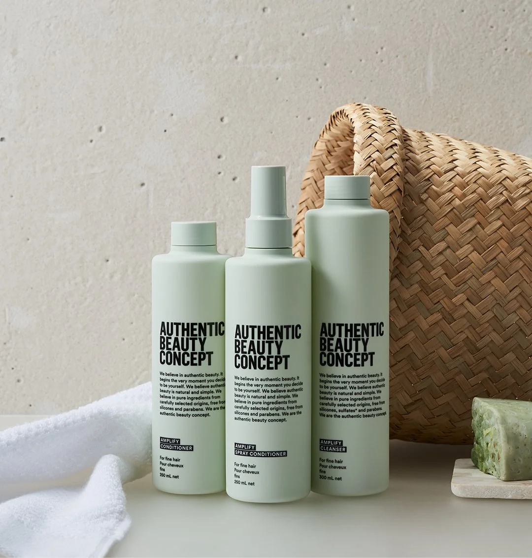 Three bottles of Authentic Beauty Concept hair care products on a white surface, with a woven basket and a green soap bar in the background.