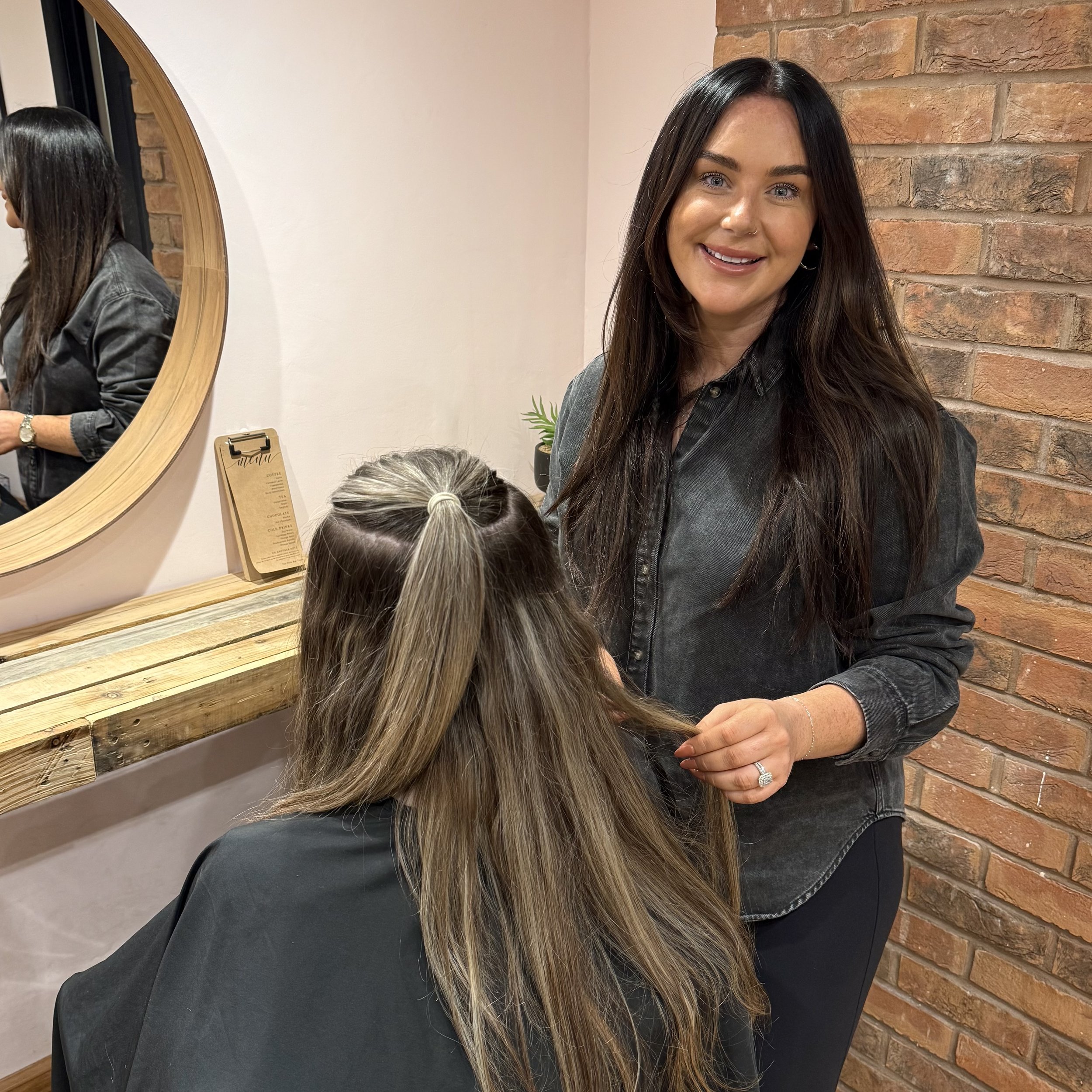 A hairstylist with long dark hair smiling and holding a section of client's hair in a salon, with a background brick wall, mirror, and a small plant.