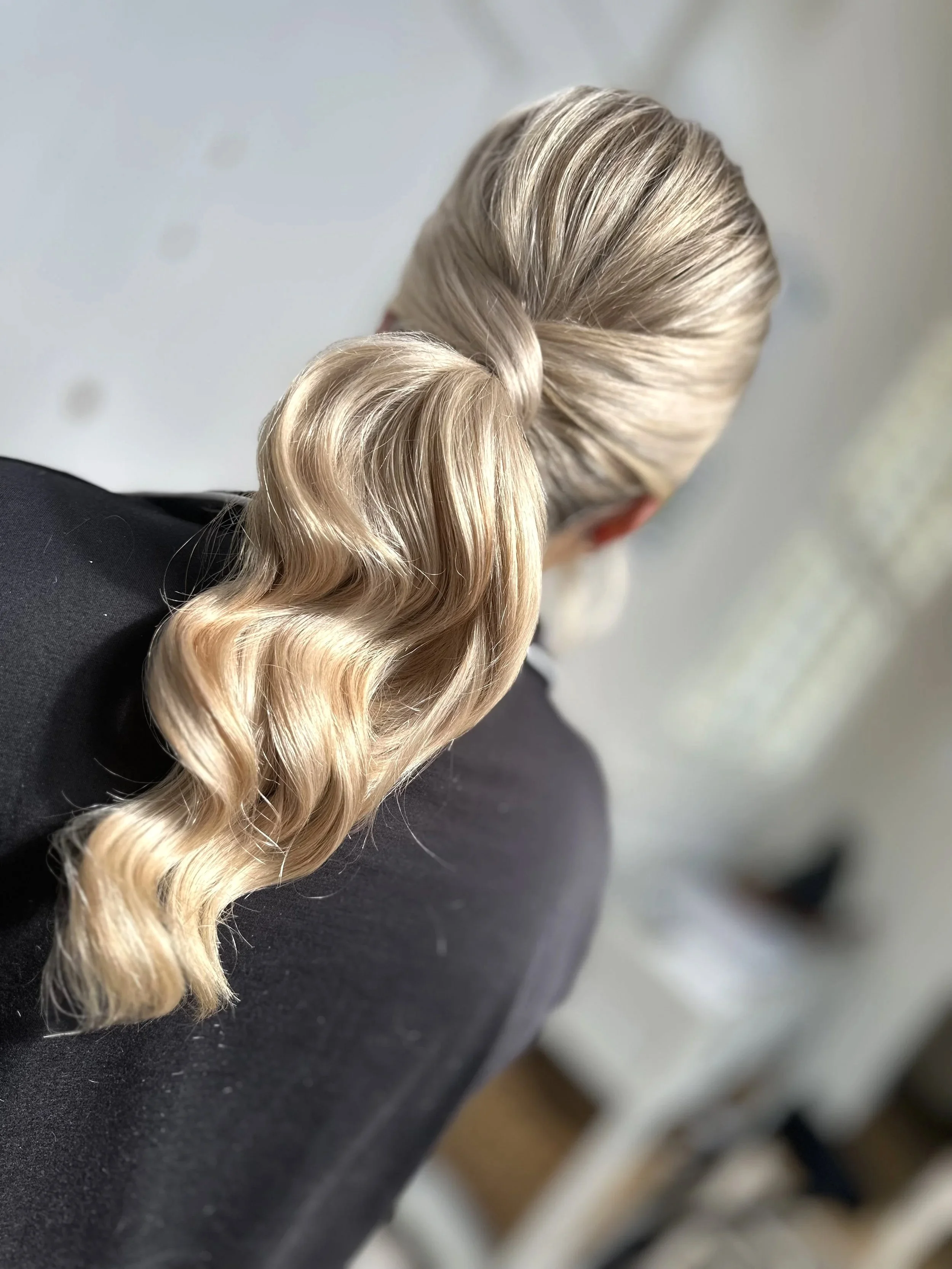 Back of a woman with styled blonde hair in loose waves, wearing a black top.