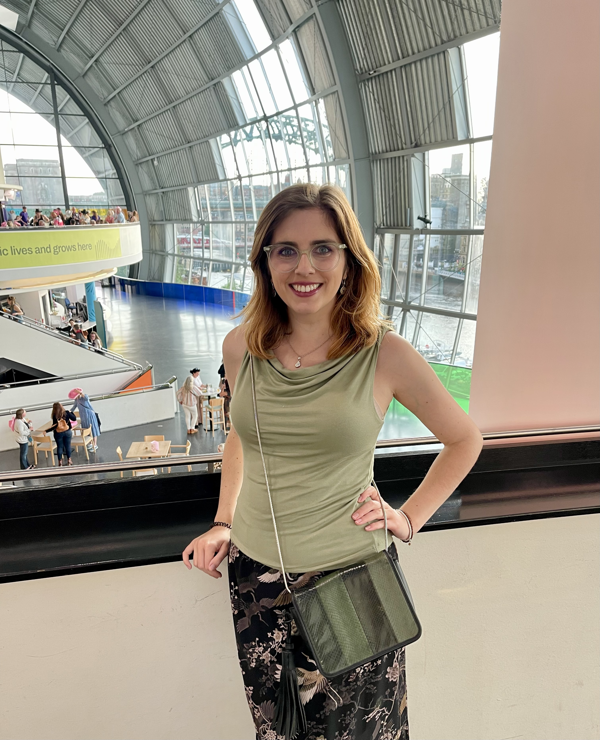A young woman with glasses, smiling, standing indoors near a railing with a large glass window behind her. She is wearing a green sleeveless top, black floral skirt, and has a small green crossbody bag. The background shows a spacious, modern building with a high arched ceiling and what appears to be a harbor or waterfront outside.