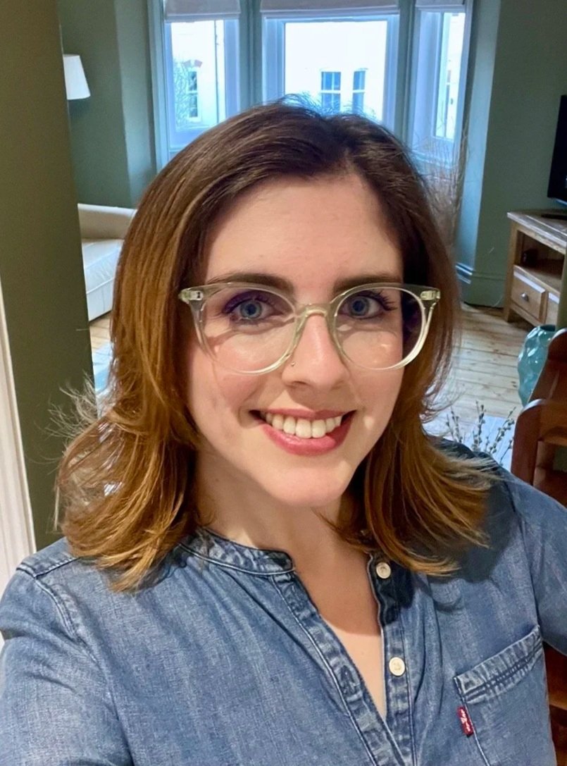 A woman with shoulder-length brown hair, glasses, and a denim shirt, smiling in a cozy living room with large windows, a couch, a TV, and hardwood floors.