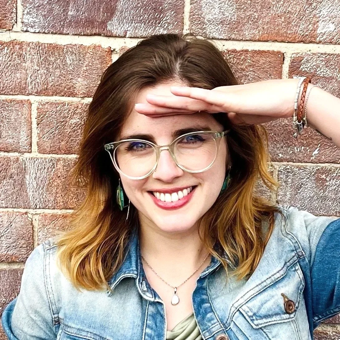 A smiling woman with glasses and earrings, wearing a denim jacket, shielding her eyes with her hand in front of a brick wall.