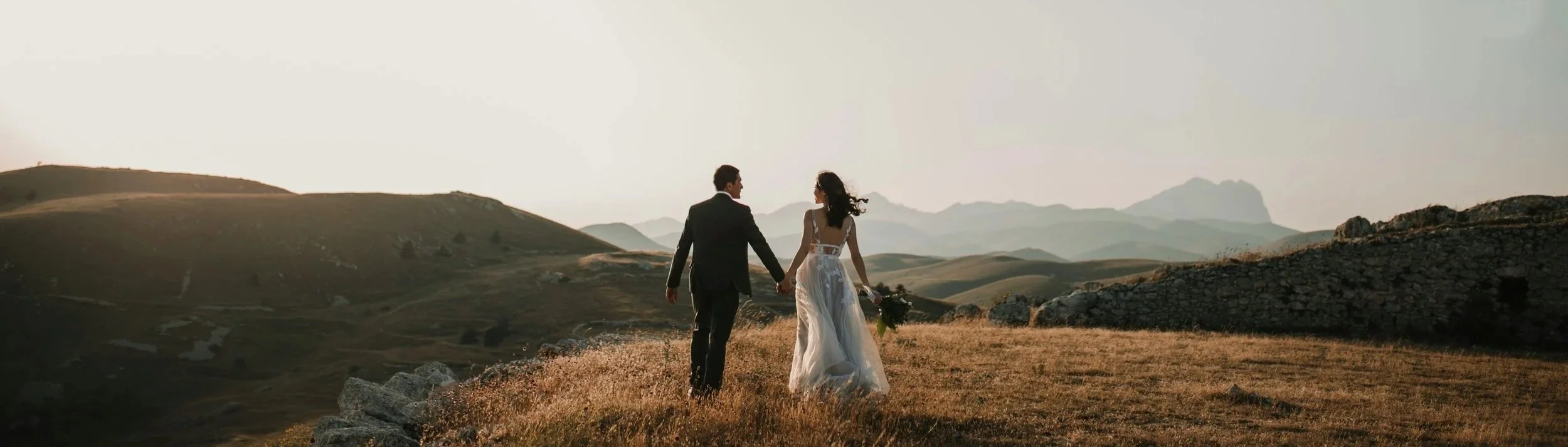 A bride and groom walking hand in hand through a grassy, hilly landscape as the sun sets, with mountains in the background.