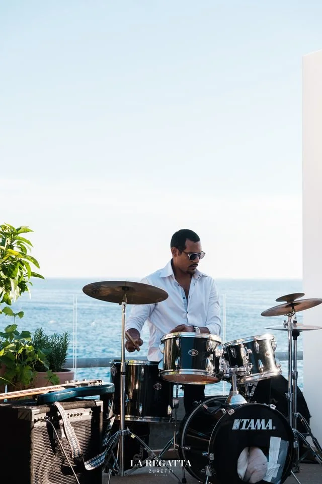 Un homme joue de la batterie sur une terrasse avec vue sur la mer, portant une chemise blanche et des lunettes de soleil.