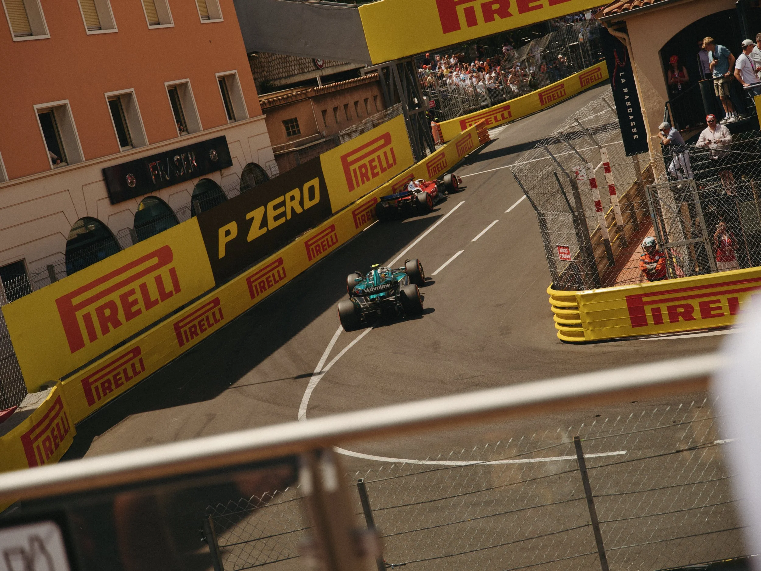 Une scène de course automobile dans une rue, avec un circuit et des barrières de sécurité. Une voiture de course est en train de tourner sur le circuit, entourée de spectateurs et de membres de l'équipe