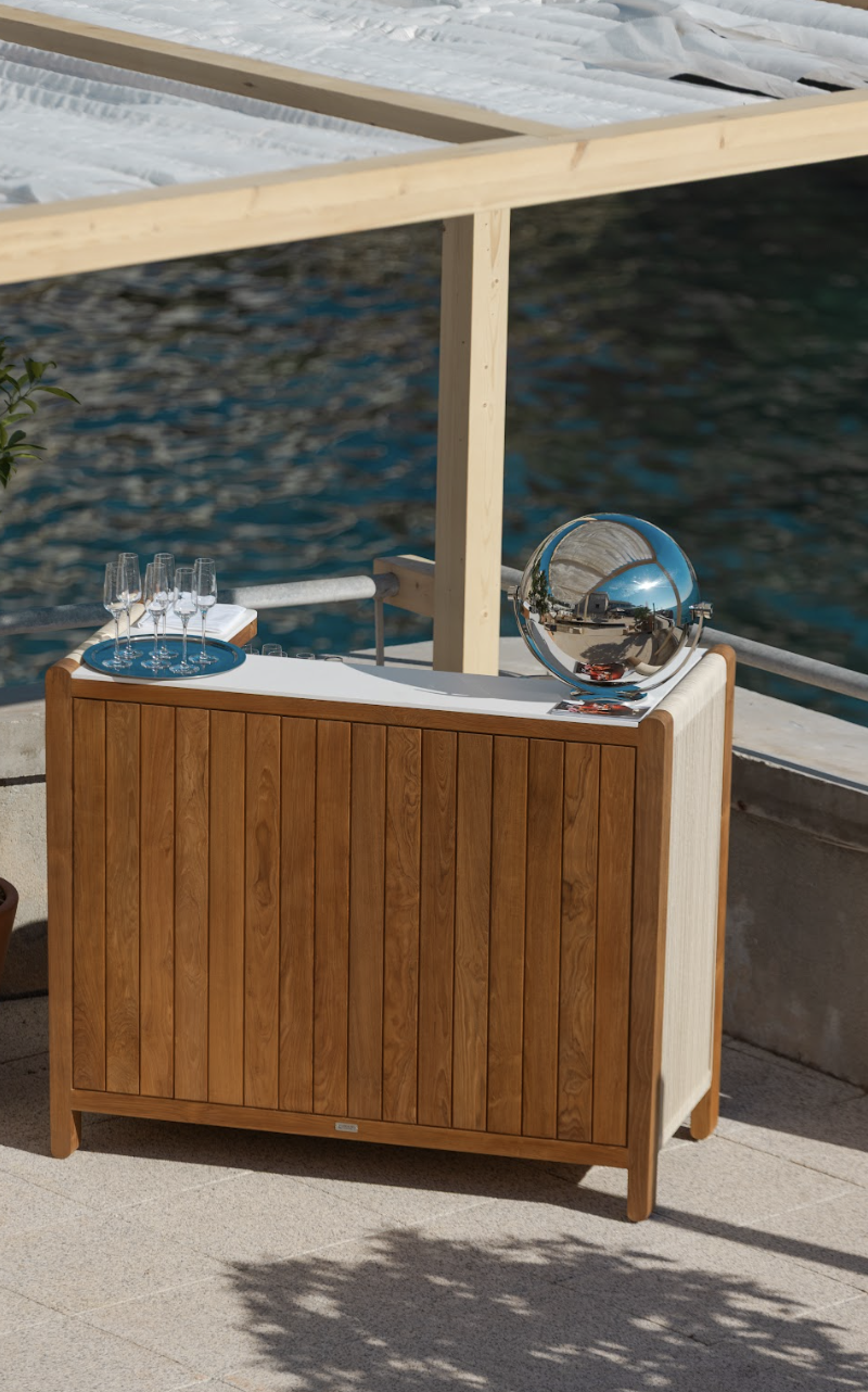 Un meuble en bois avec des verres à champagne et une boule en métal réfléchissante, situé à côté de l'eau en extérieur.
