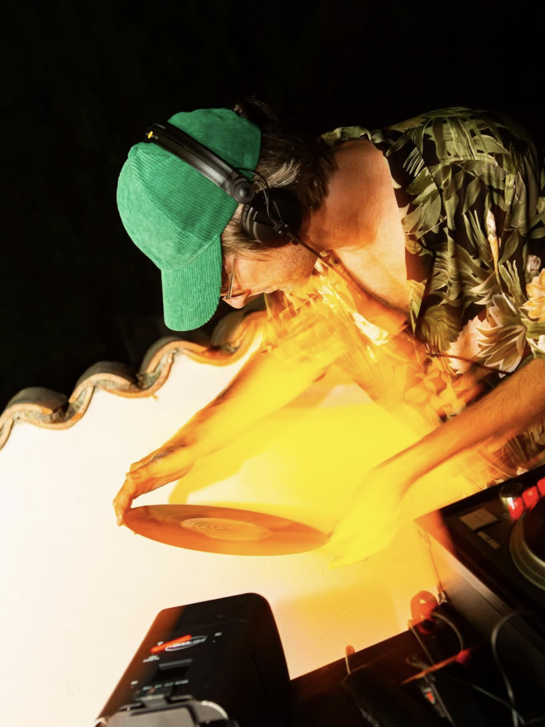 Un homme portant une chemise à motif floral, un casque audio vert et une casquette verte, qui manipule une boule de laine orange sous une lumière orange
