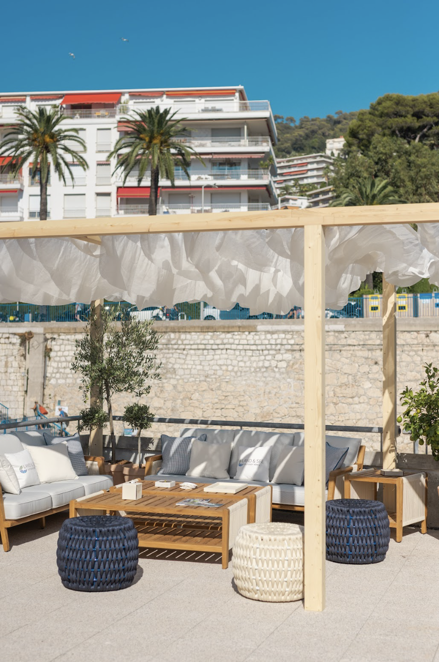 Espace extérieur avec des sièges en bois, coussins et poufs tressés sous une pergola, en face d'un mur en pierre et au-delà de palmiers et d'un bâtiment résidentiel blanc avec balcons et volet rouge, contre un ciel bleu clair.