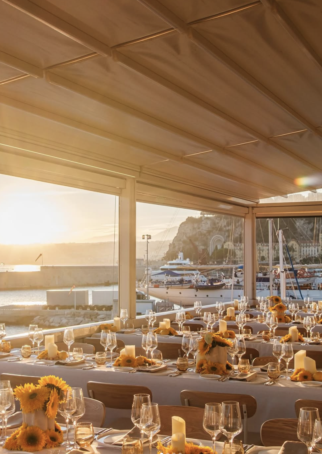 Salle de banquet décorée avec des tables avec des nappes blanches, des bougies et des bouquets de tournesols, avec une vue sur un port de plaisance au coucher du soleil.