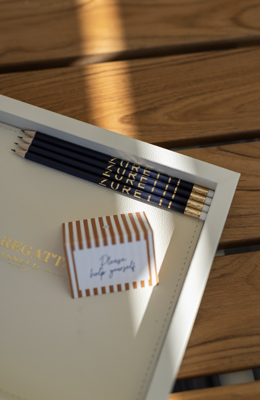 Cahier ou carnet blanc avec des crayons noirs et dorés portant le nom ZURETI, à côté une petite carte avec des rayures orange et blanches et le message 'Dear best yourself'