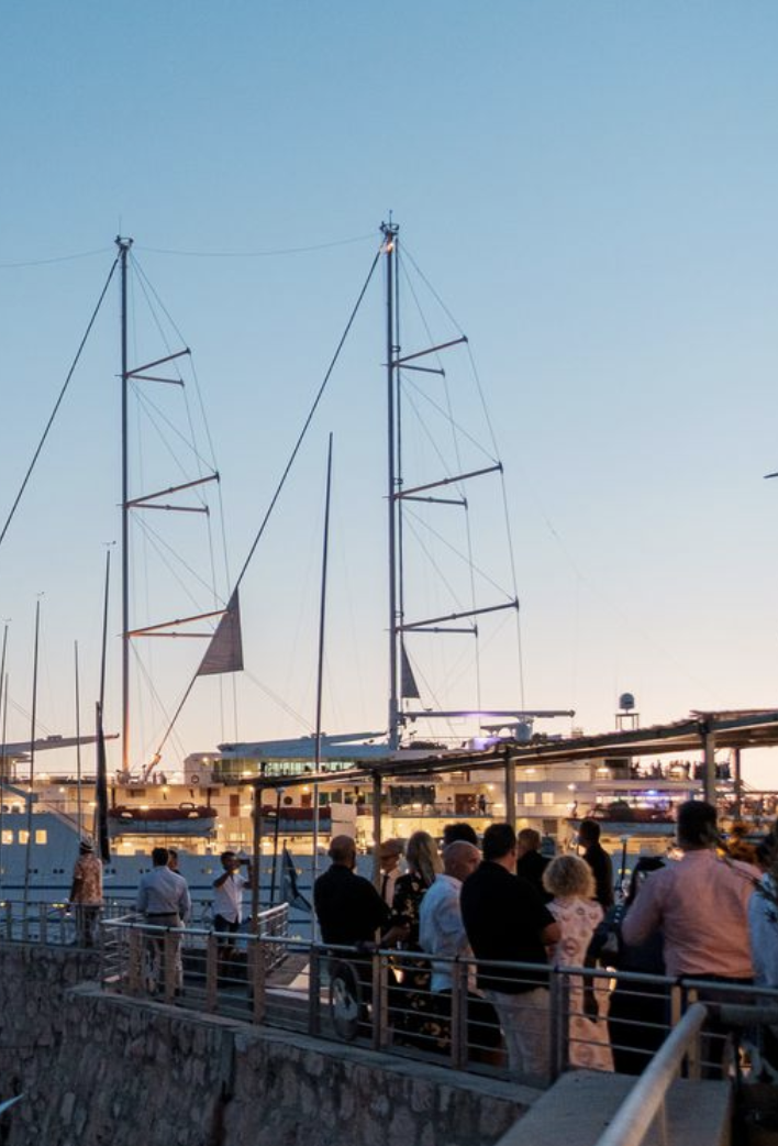 Gens debout sur une promenade au bord de l'eau, avec des voiliers amarrés en arrière-plan lors du crépuscule.
