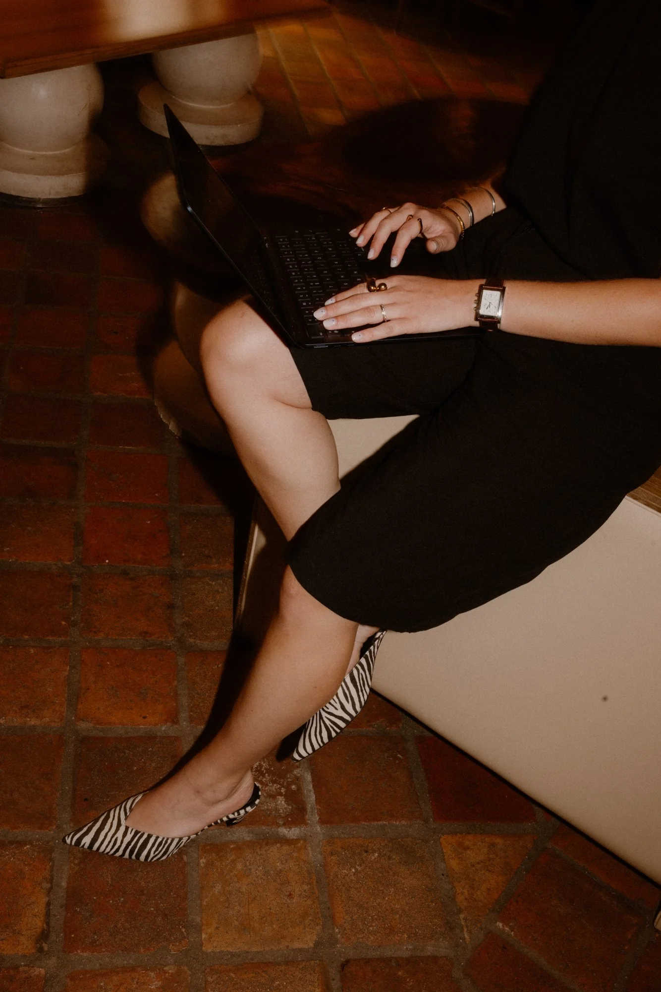 Une personne assise sur un banc en cuir, utilisant un ordinateur portable, portant une robe noire, des chaussures à motif zèbre, une montre et plusieurs bracelets.