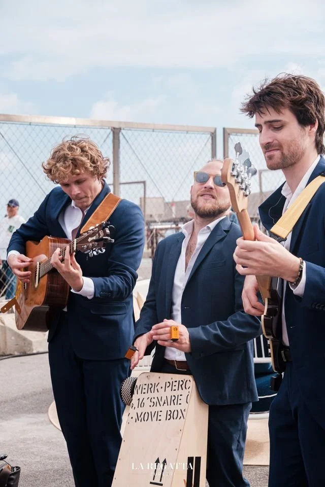 Trois hommes en costume jouent de la musique avec des guitares, un d'eux porte des lunettes de soleil, dans un espace en plein air encadré par une barrière en métal.
