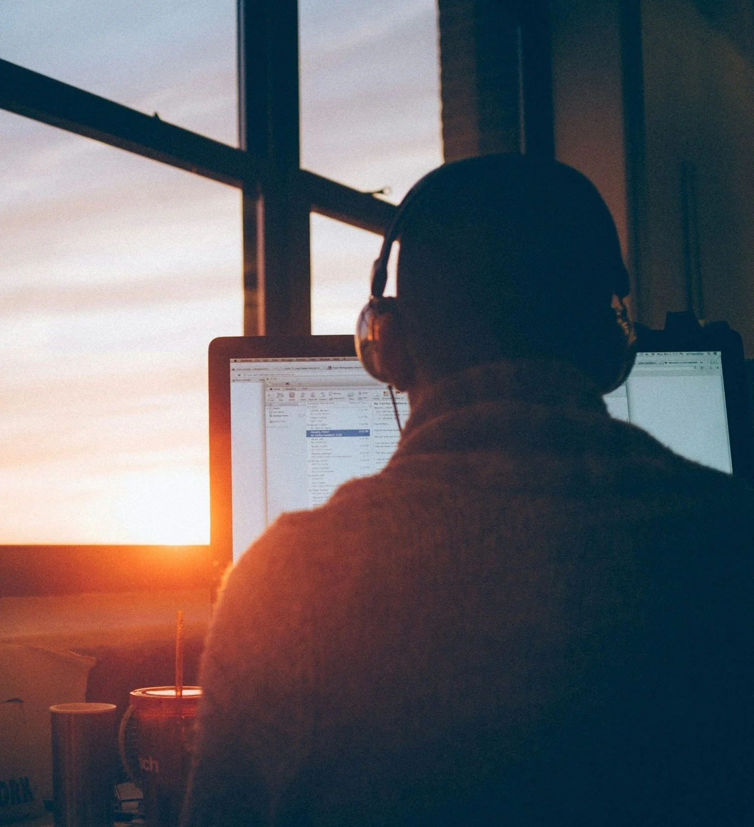 Person wearing headphones sitting at a desk with a computer, backlit by sunset through a window. Screen displays an email application.