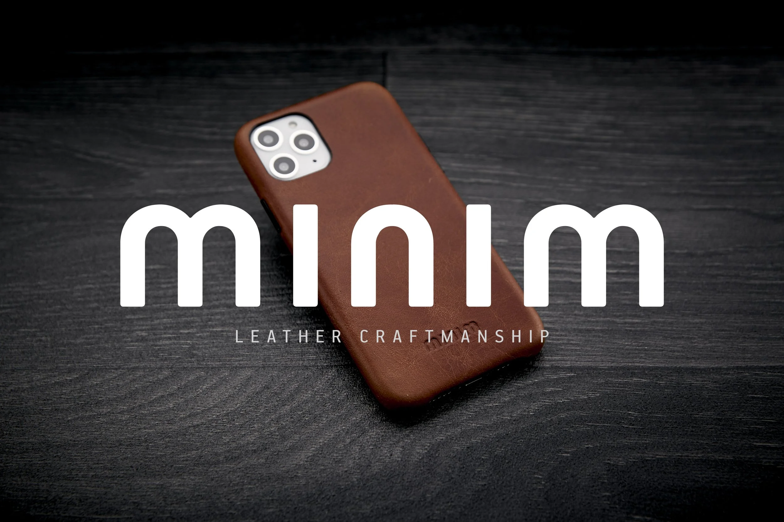 Bruine leren telefoonhoes op zwart hout met tekst 'minim leather craftsmanship'.
