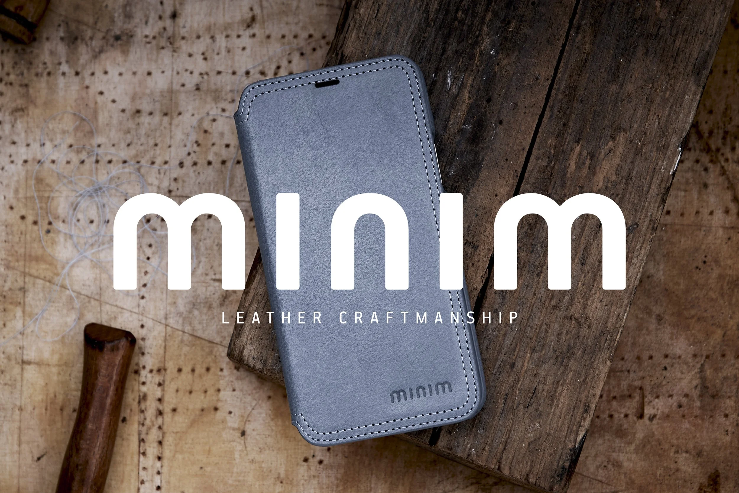 Grijze leren telefoonhoes met Minim-logo, geplaatst op een houten oppervlak, met de tekst "Minim Leather Craftmanship" eroverheen.