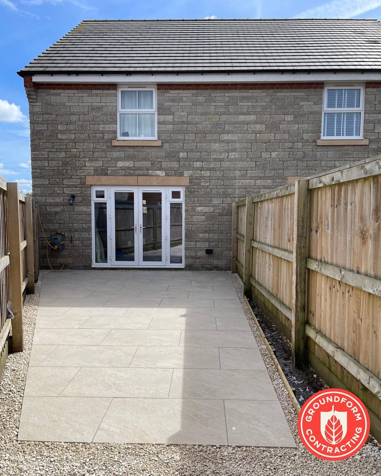 Backyard patio with new beige tiles surrounded by gravel, enclosed by a wooden fence, with a brick house and double glass door leading inside. There are two windows above the patio, and a small outdoor light fixture on the wall.