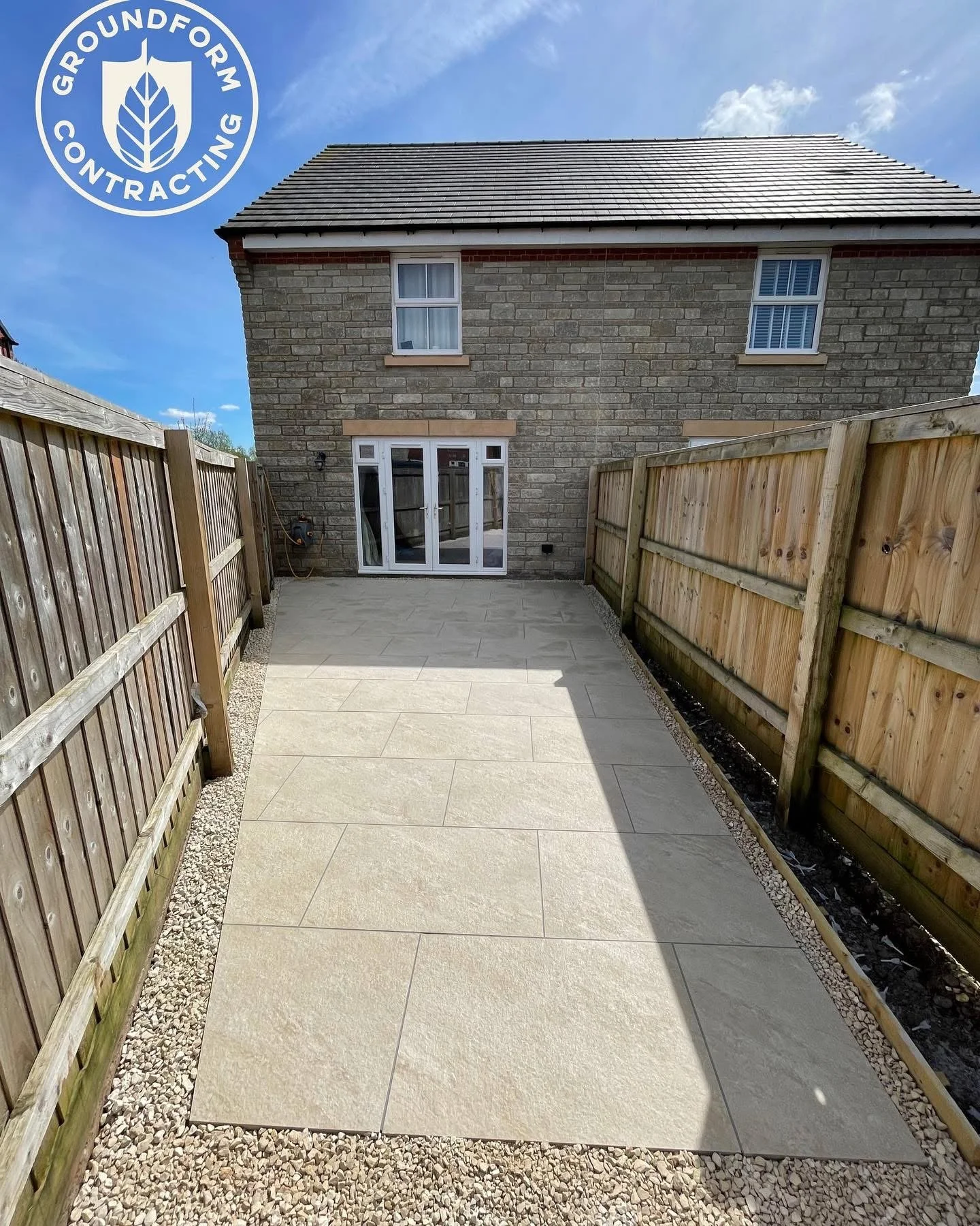Backyard patio with newly laid large beige tiles, bordered by small white pebbles, enclosed by wooden fences, and a brick house with a sliding glass door and two windows in the background.