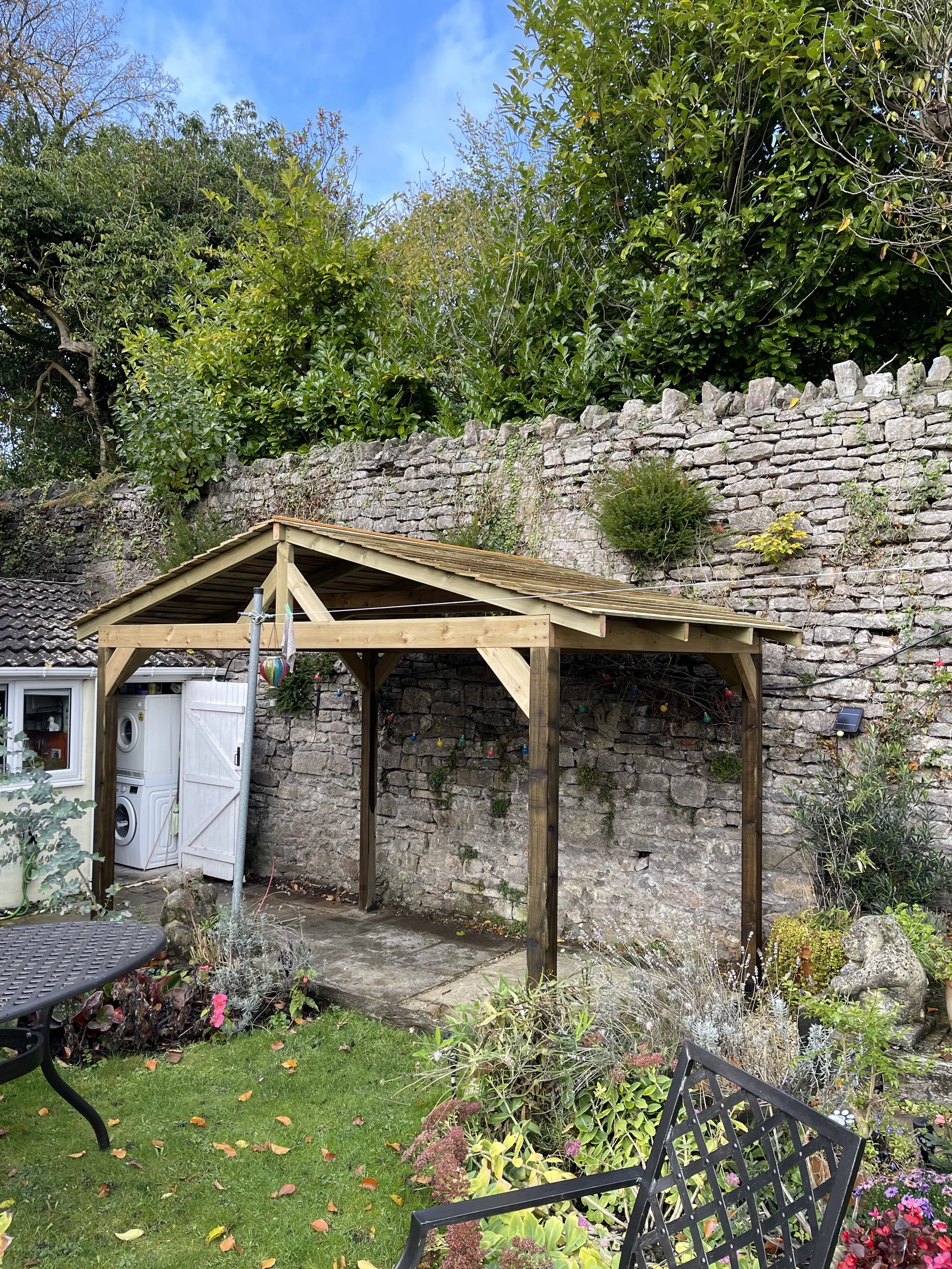 A backyard garden with a small wooden shelter under construction, surrounded by plants, flowers, and garden furniture, with a stone wall and large trees in the background.