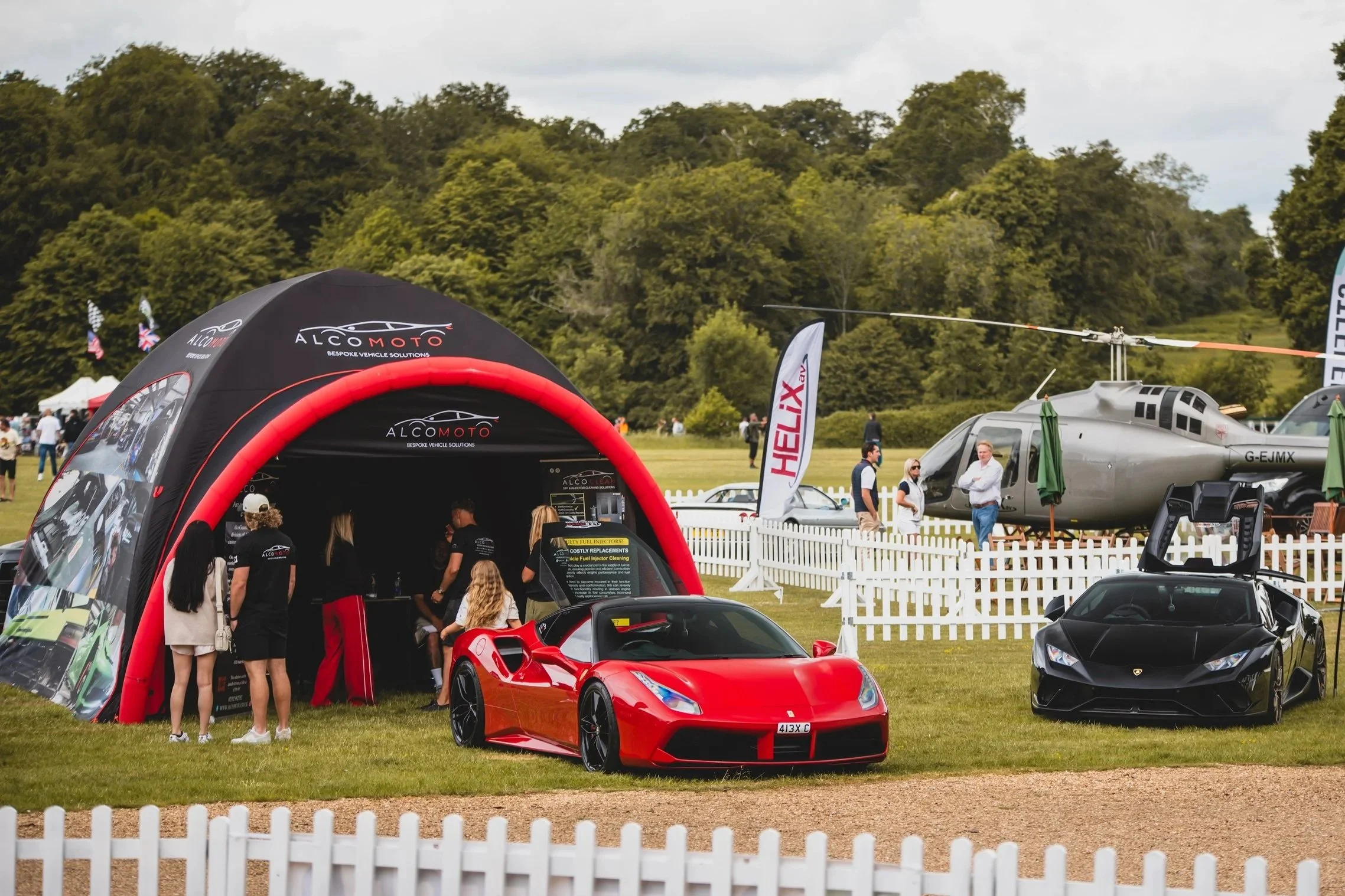 Red Ferrari parked on grass behind white picket fencing at an outdoor event with tents, a helicopter, and people in the background.